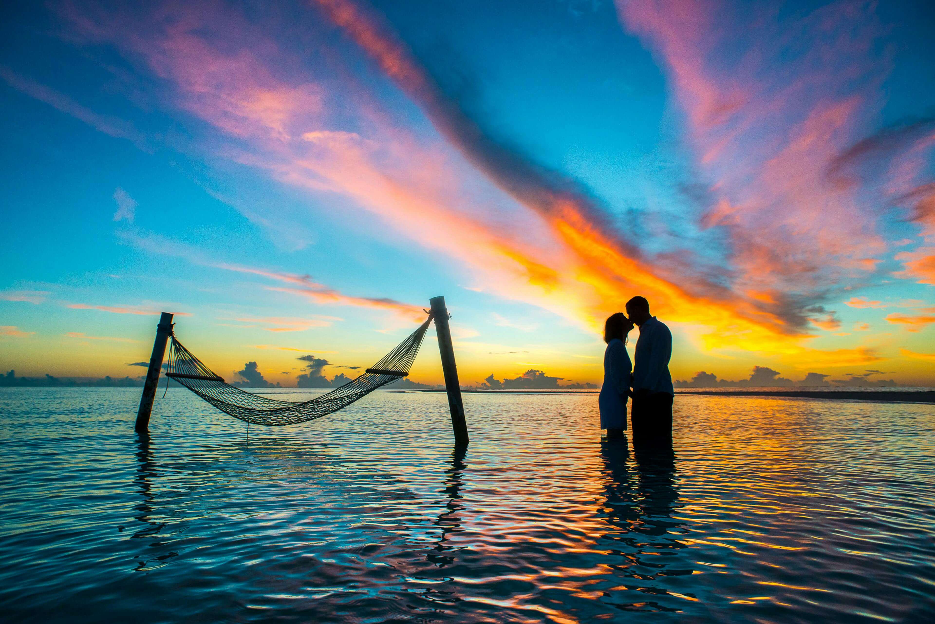A romantic couple shares a kiss at sunset with vibrant skies over the Maldives beach.