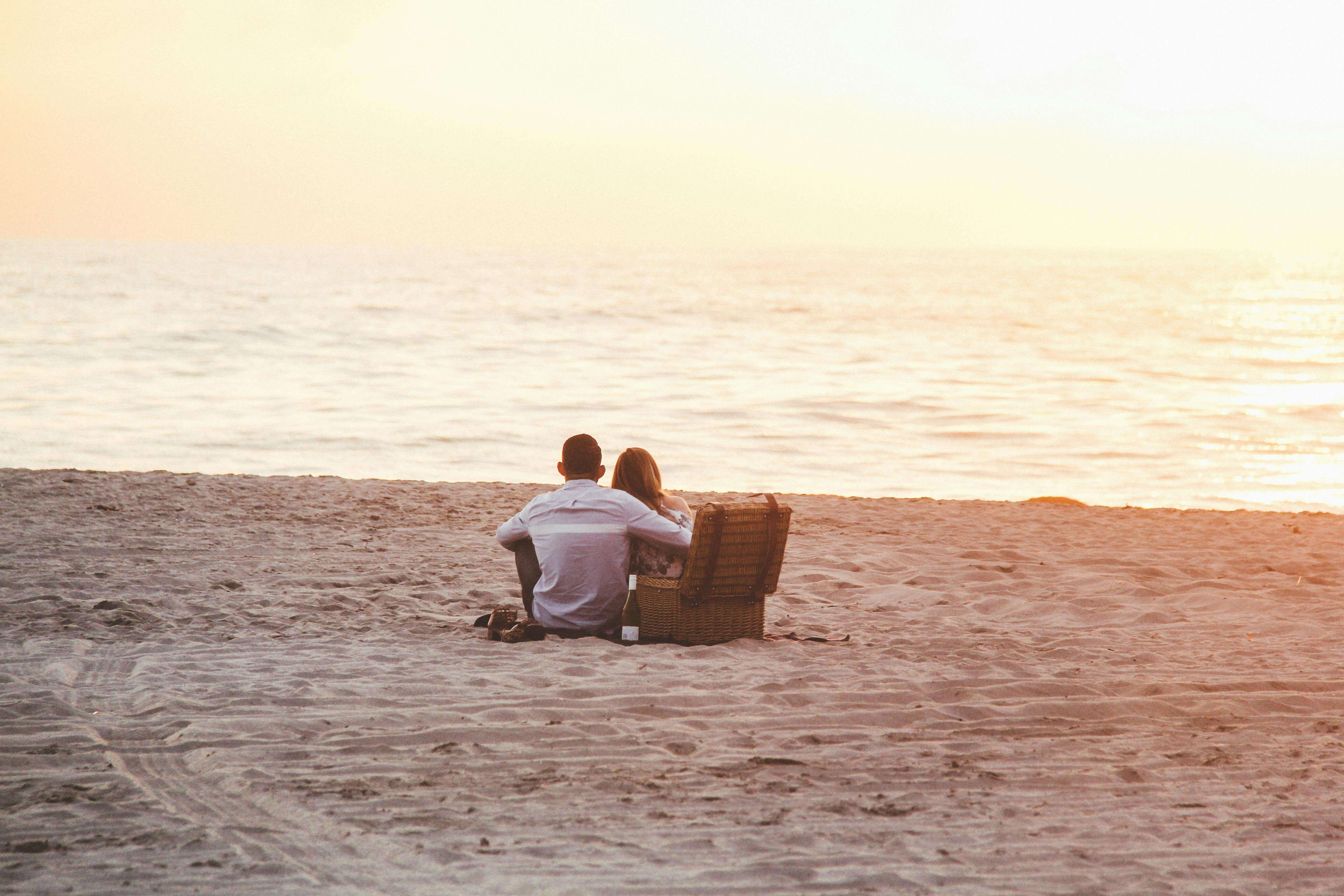 A couple sitting on a sandy beach, enjoying a romantic sunset picnic by the ocean shore.
