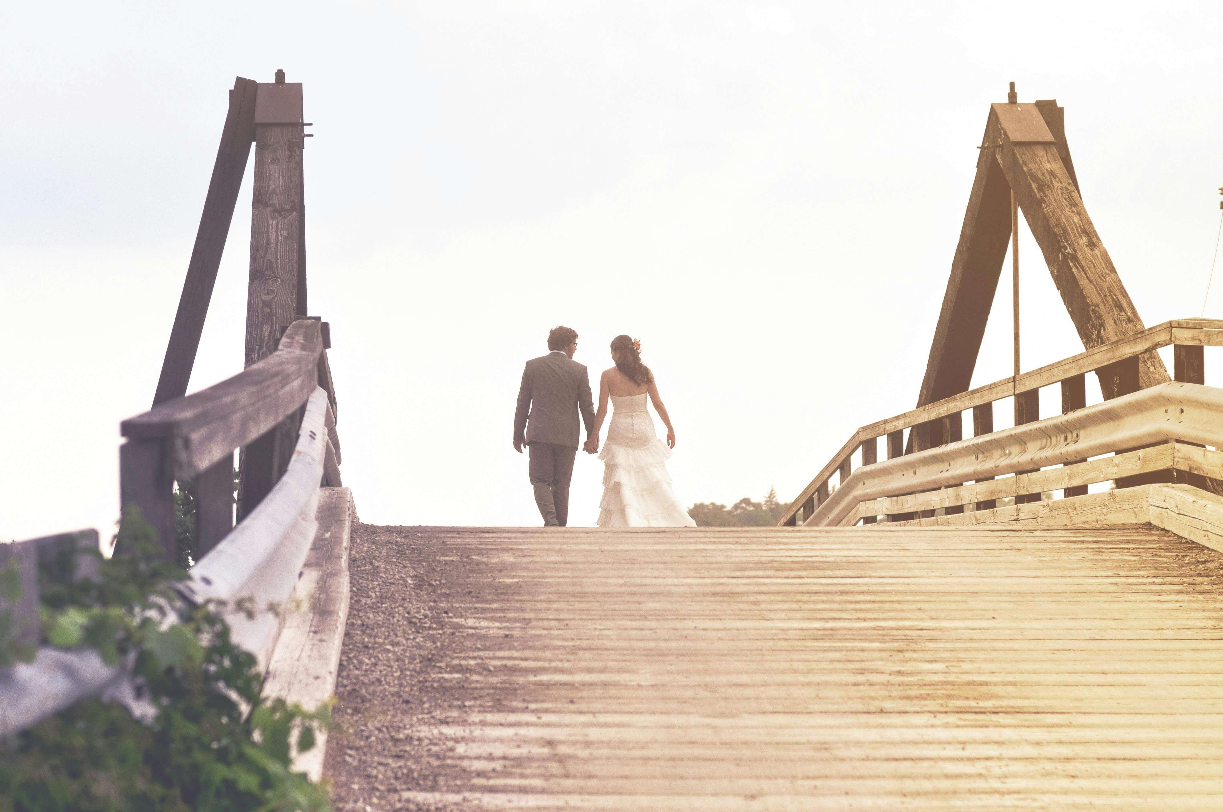 A bride and groom holding hands walking on a wooden bridge during sunset, symbolizing love and togetherness.