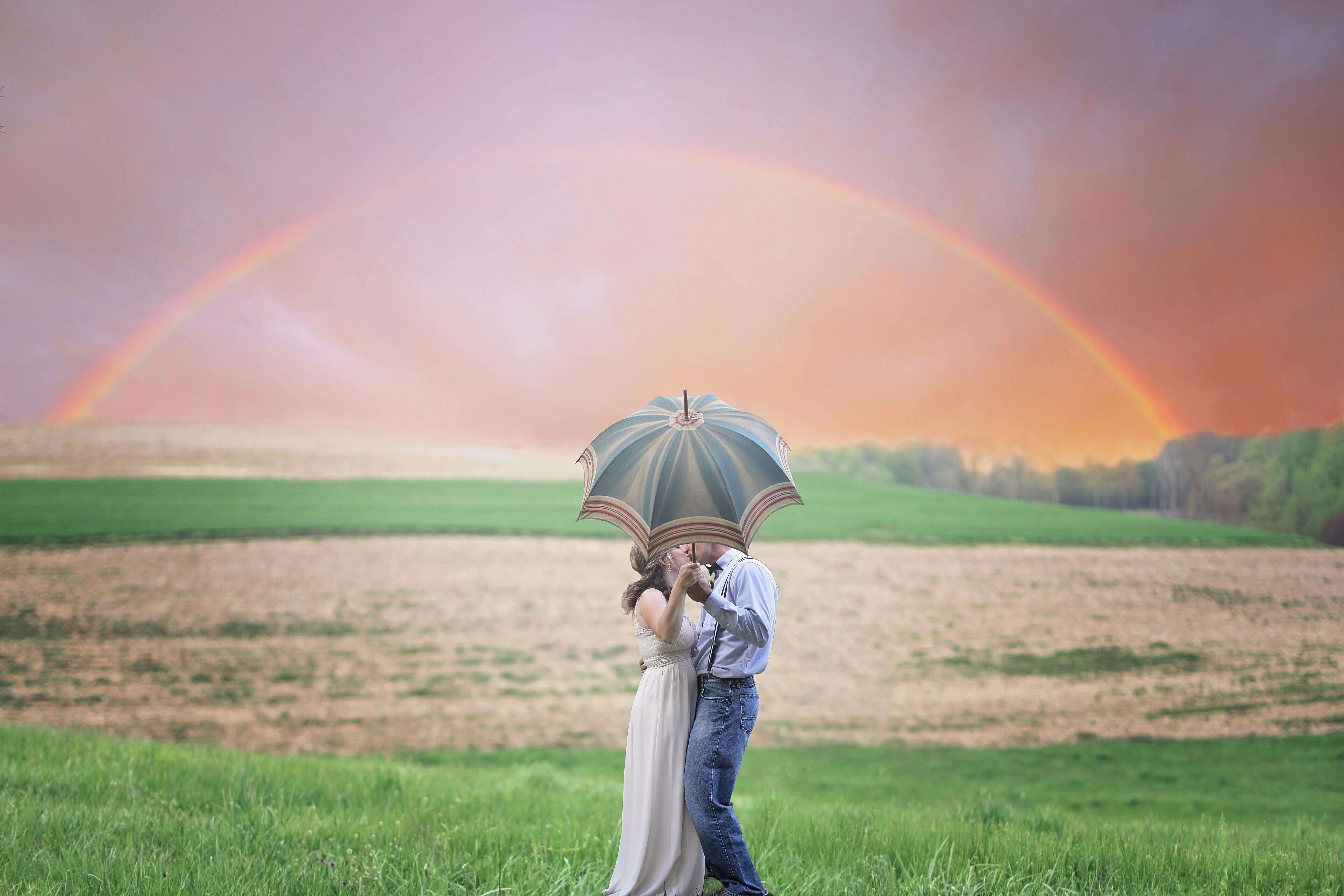 A couple shares a kiss under an umbrella in a scenic rural landscape with a rainbow.