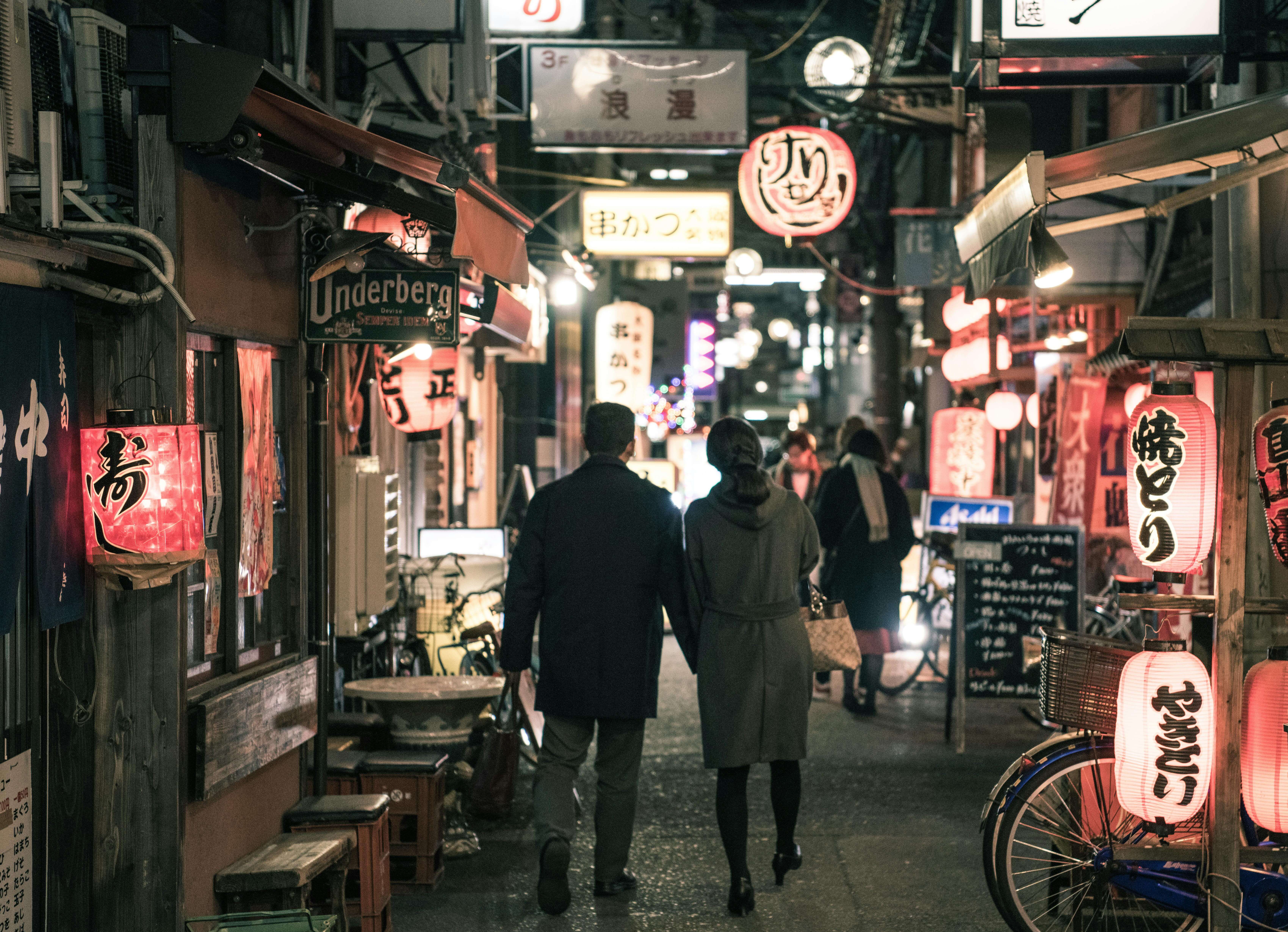 Couple strolling through Osaka's bustling alley at night, surrounded by glowing lanterns and shop signs.