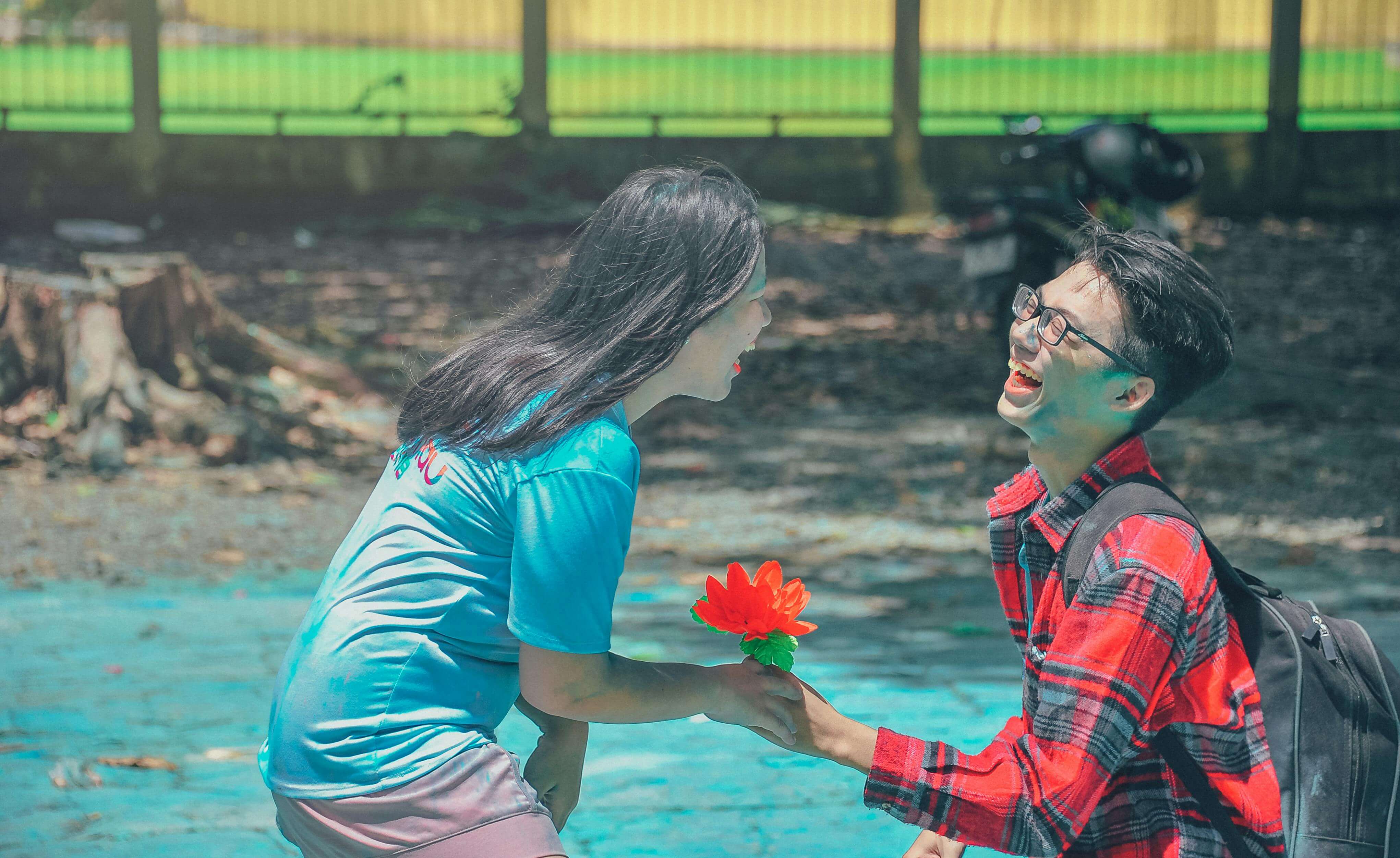 A young couple laughing and sharing a flower in a sunlit outdoor park setting.