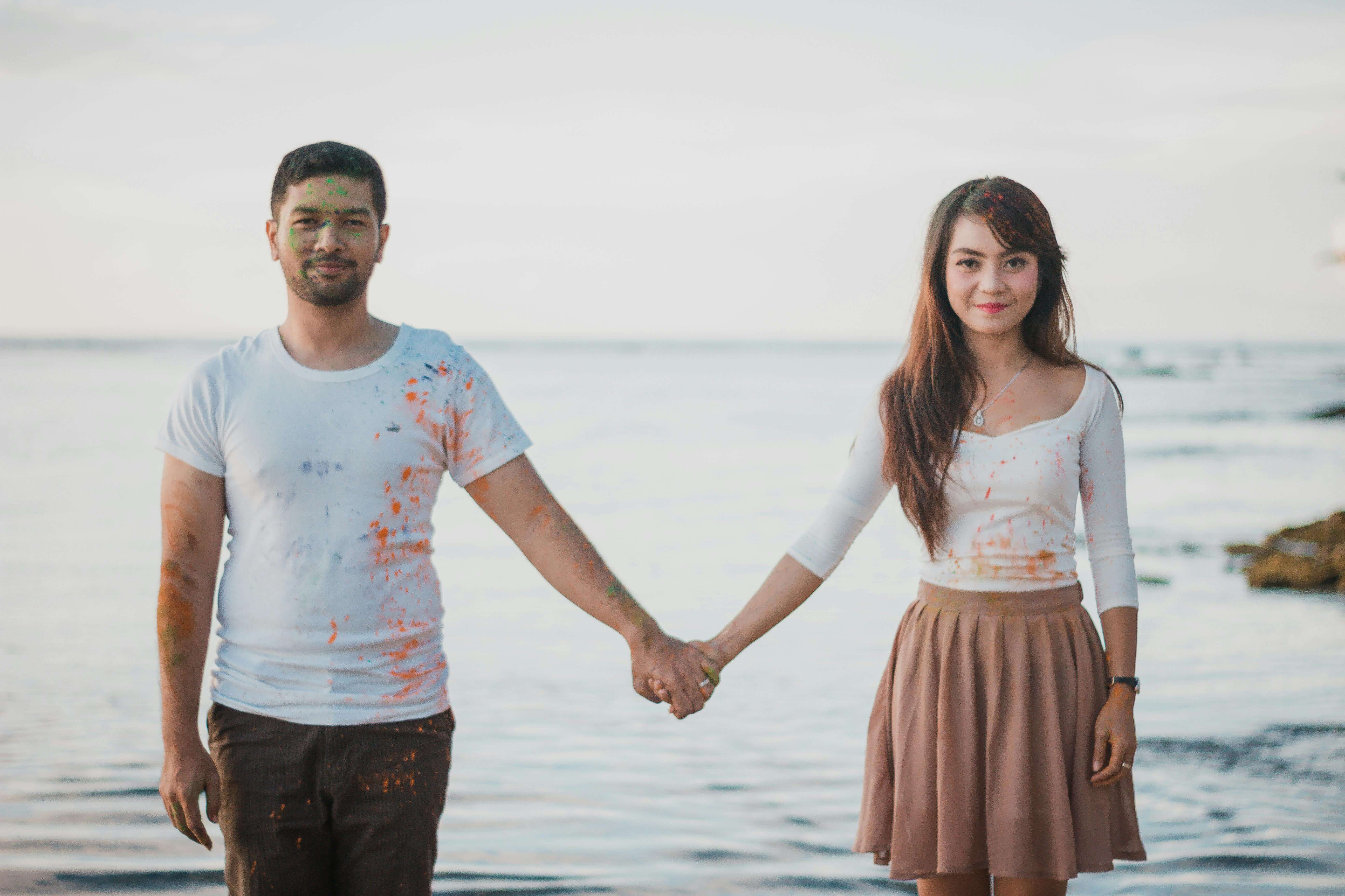 A cheerful couple holding hands by the sea, enjoying a playful day with paint splashes.