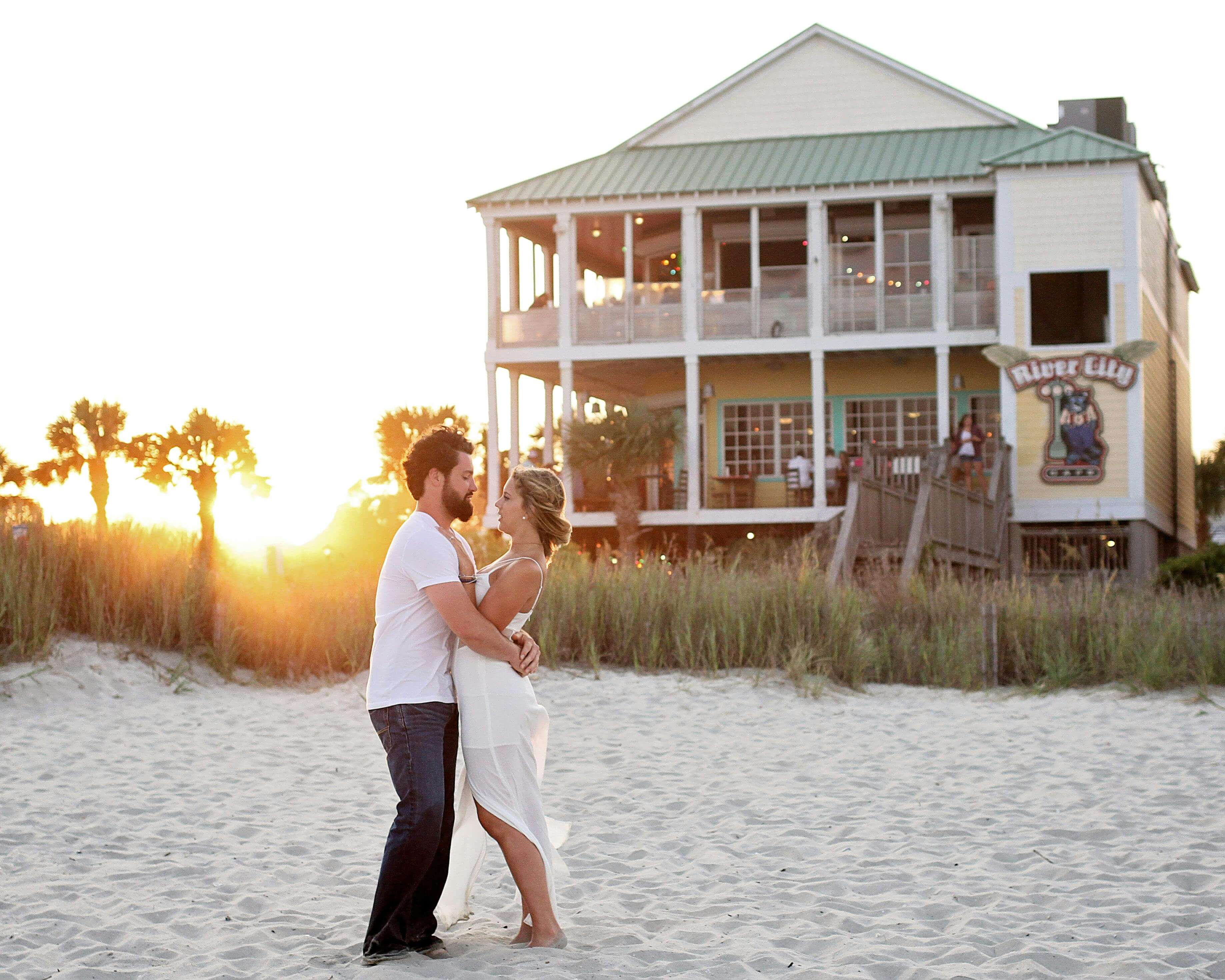 A couple embraces and dances joyfully on a sandy beach with a scenic beachfront house in the background during sunset.
