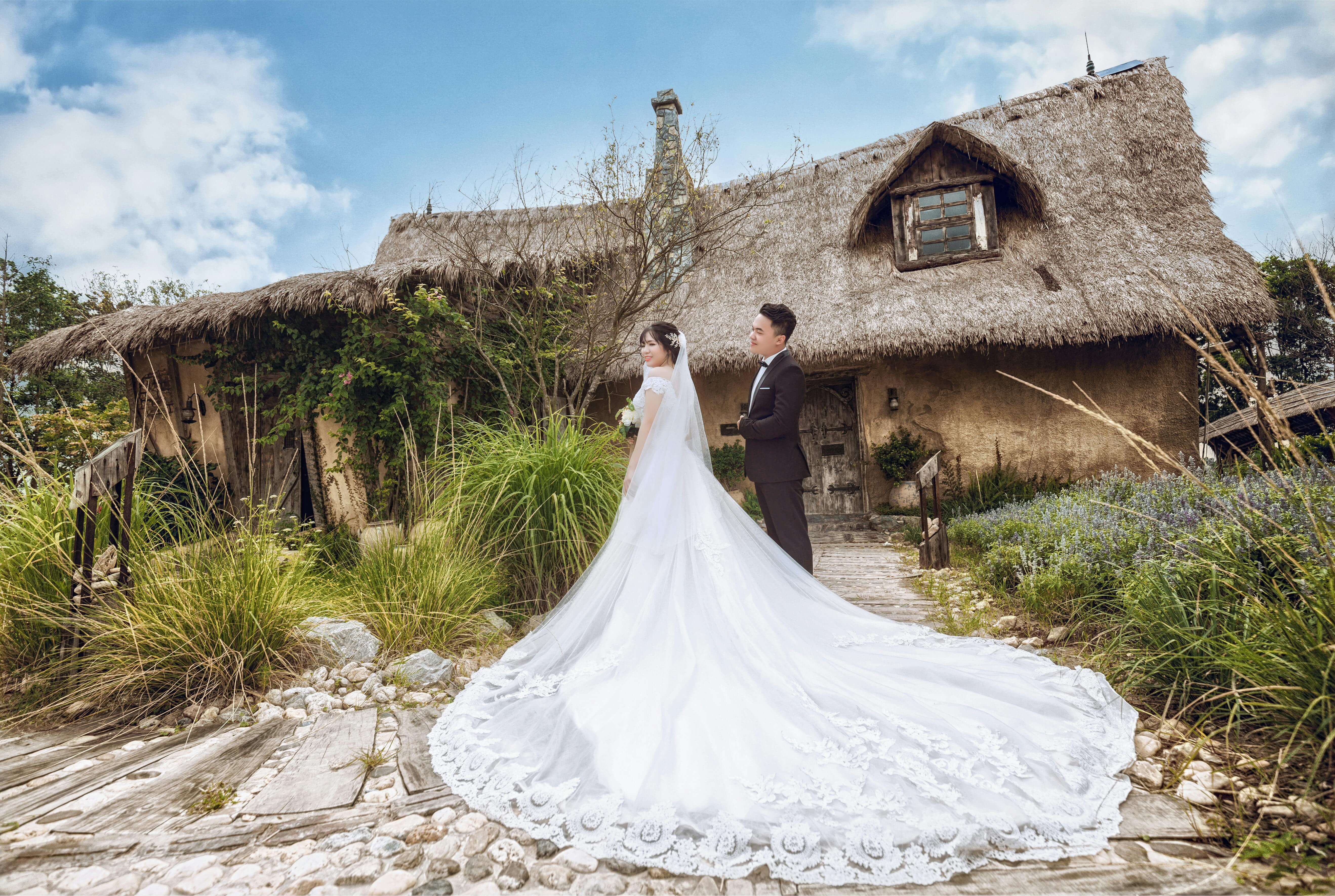 Bride and groom pose in elegant attire at a rustic outdoor location with a thatched cottage backdrop.