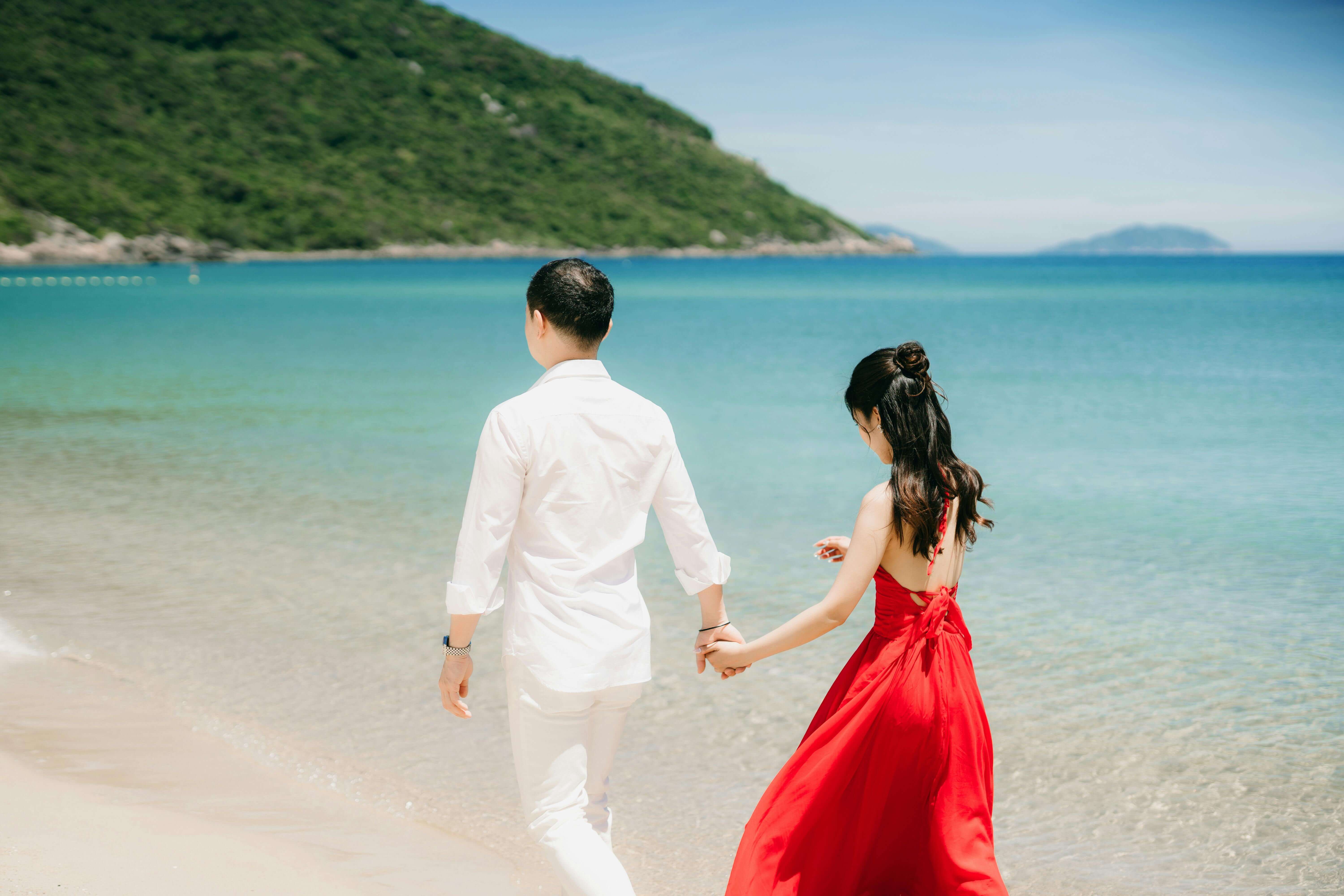 Asian couple holding hands and walking by the ocean on a sunny day, showcasing love and romance.