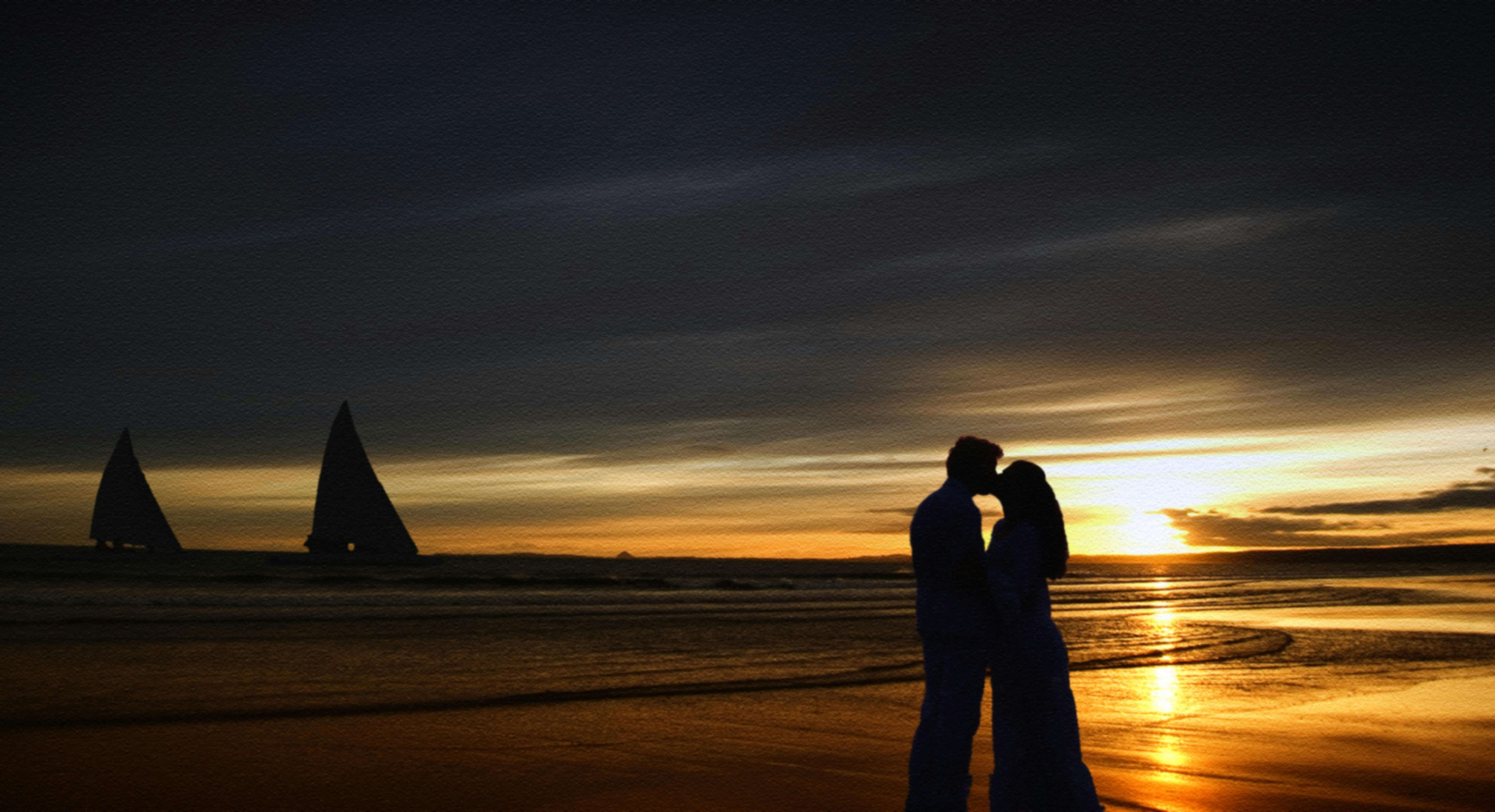 Couple embracing at the beach during a romantic sunset with sailboats on the horizon.