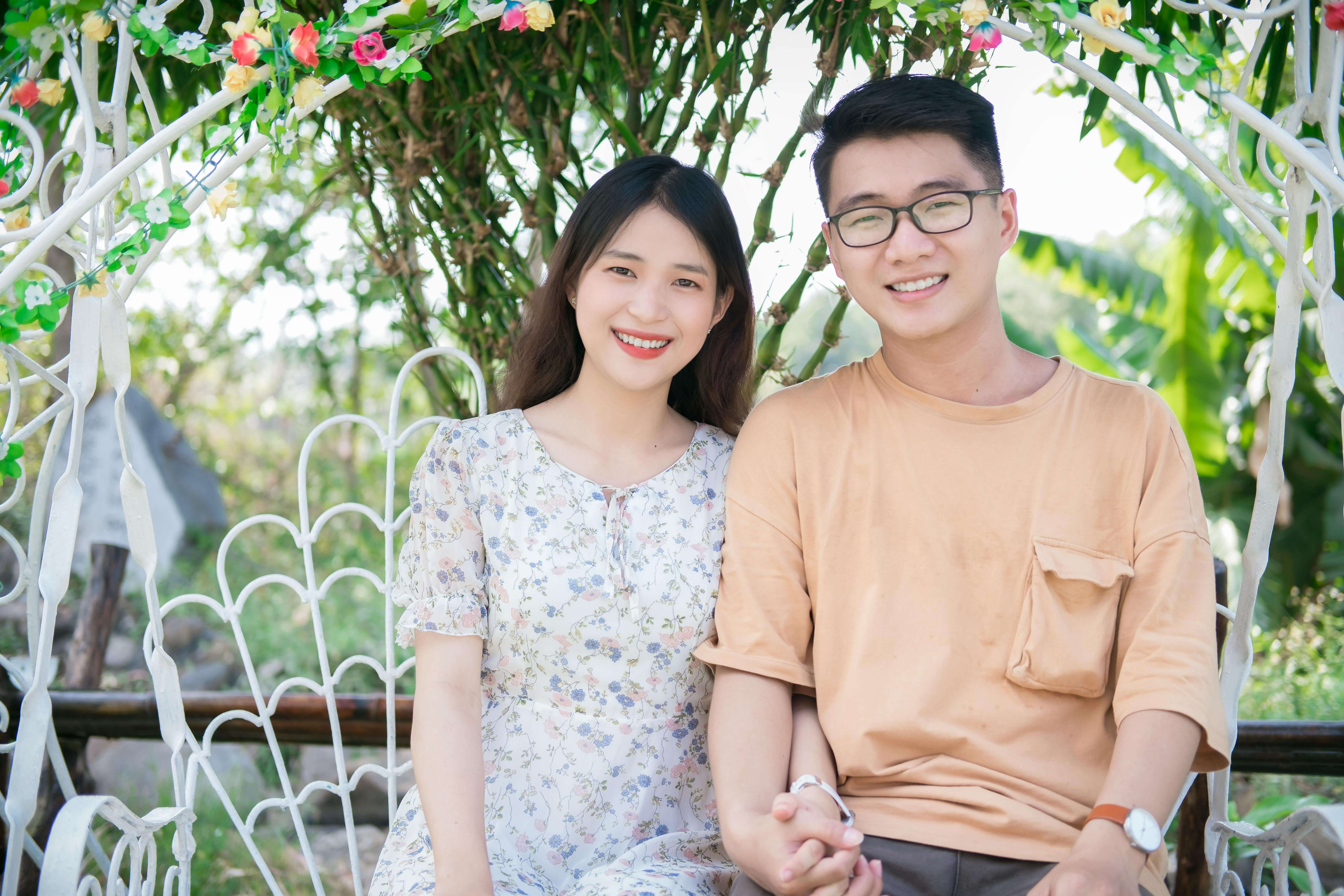 Smiling Asian couple sitting on a swing, holding hands in a lush garden.
