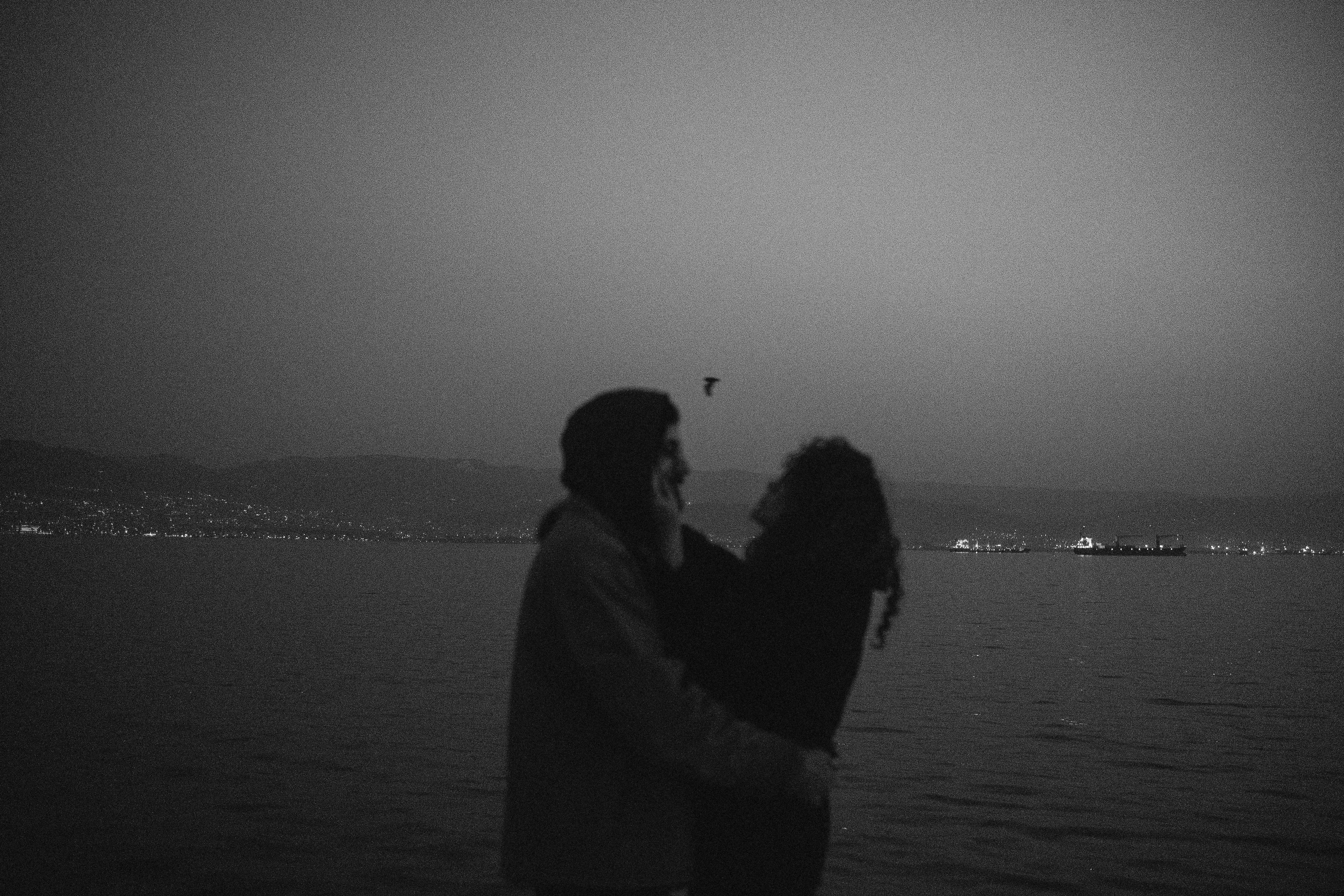 A couple embraces by the water at twilight in Ä°zmit, Kocaeli, Turkey, in a romantic black and white photo.