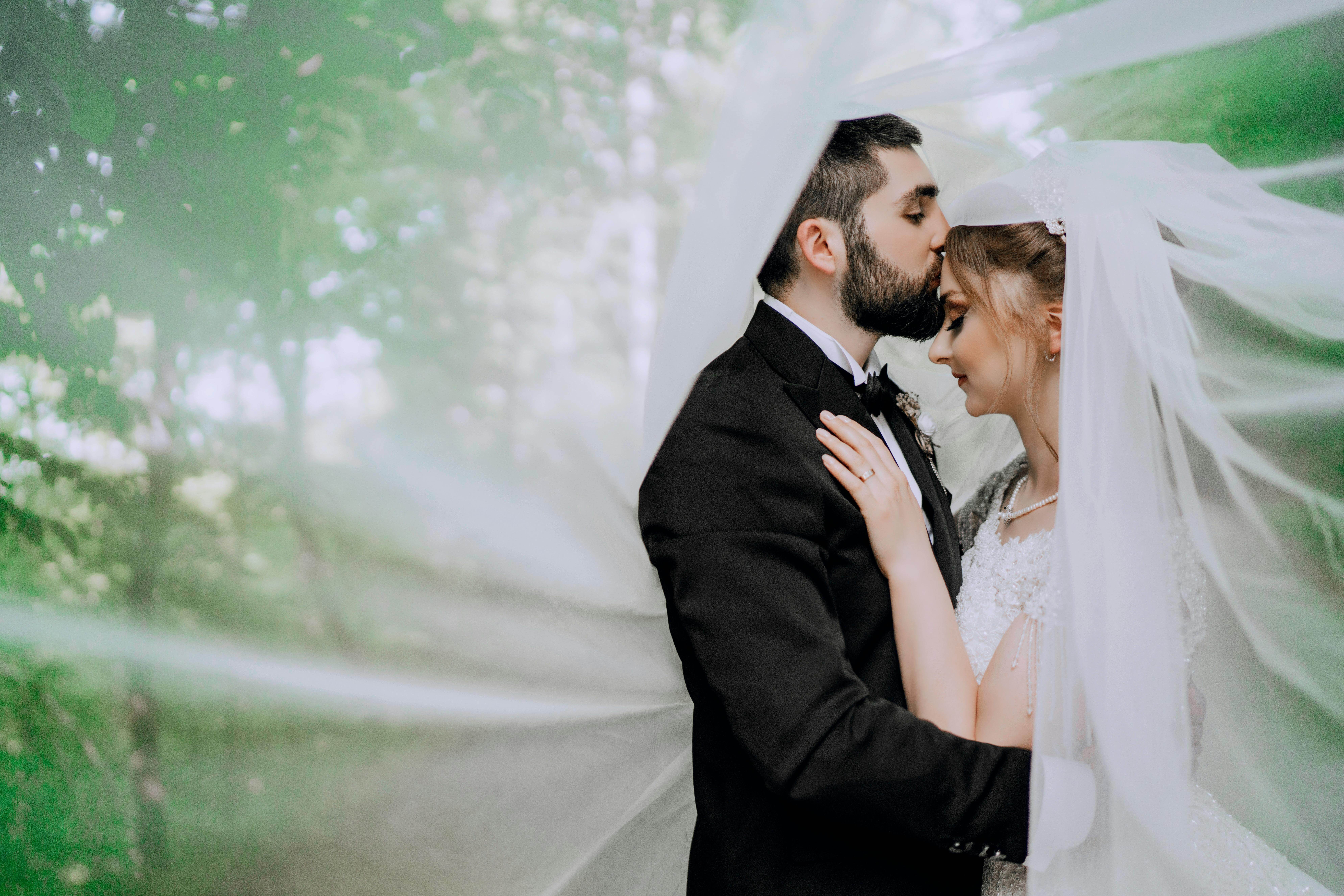 A couple in wedding attire embraces tenderly beneath a bridal veil outdoors.