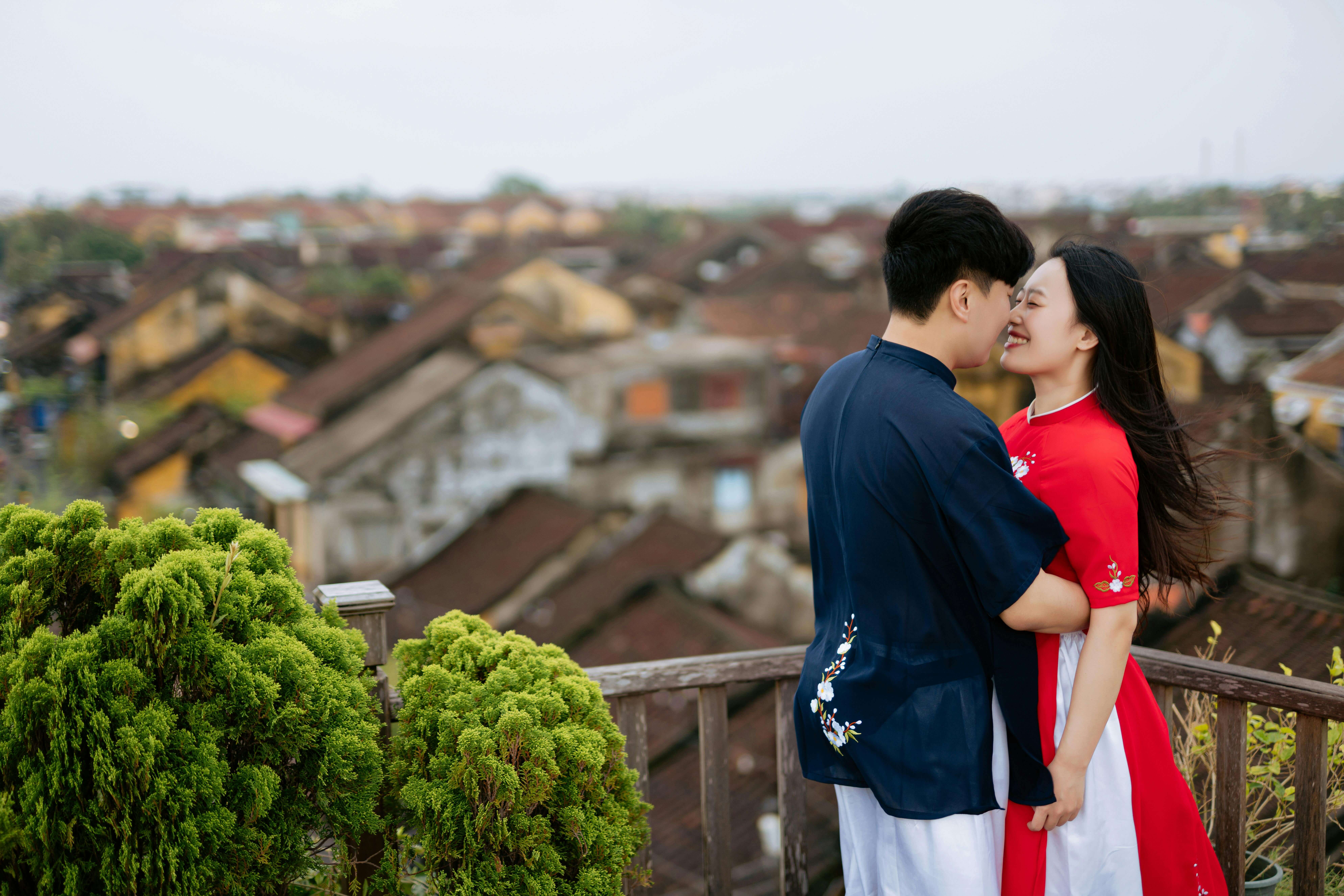 Asian couple in traditional attire sharing an intimate moment on a view deck overlooking rooftops.