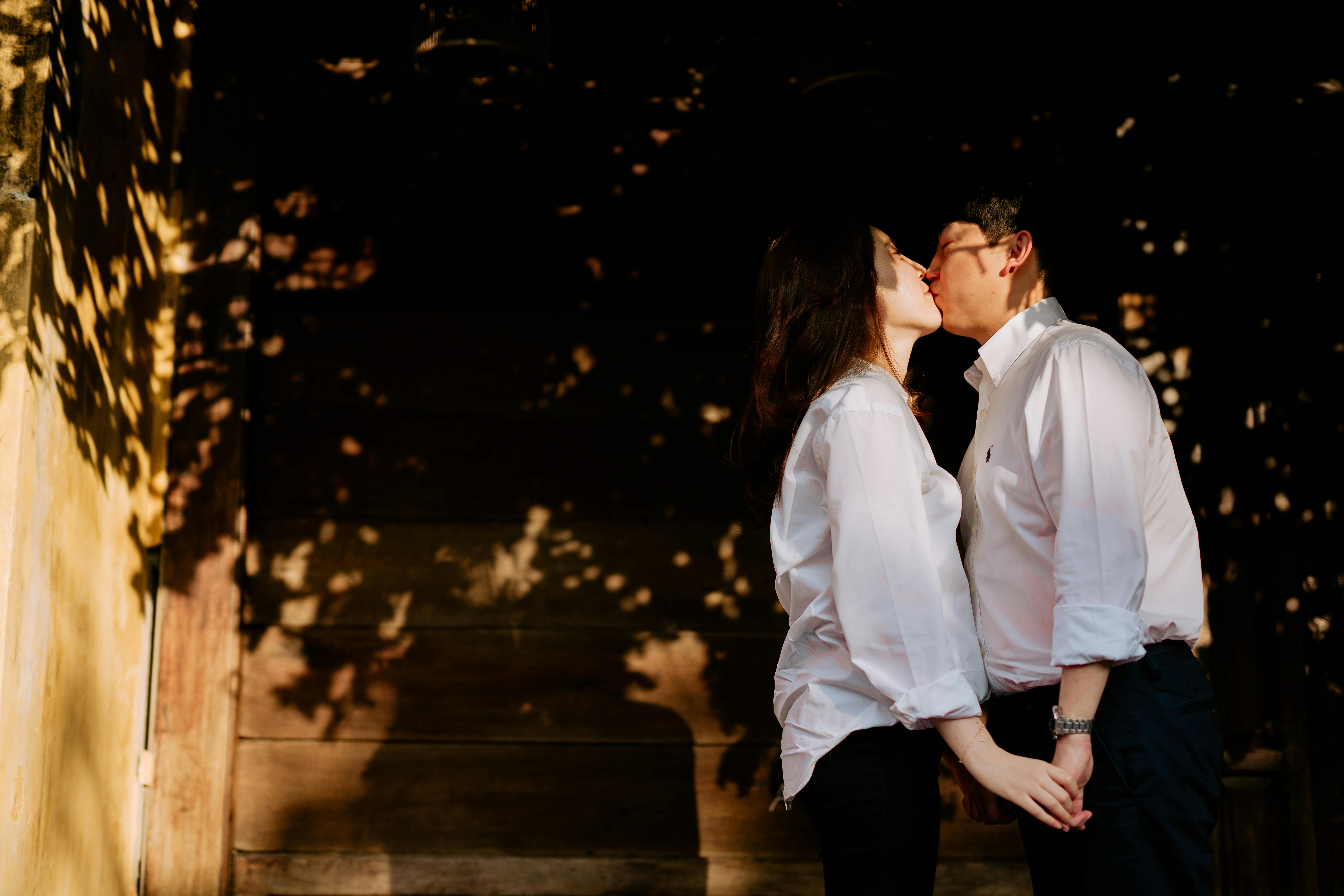 A loving couple shares a tender kiss among soft shadows outdoors.