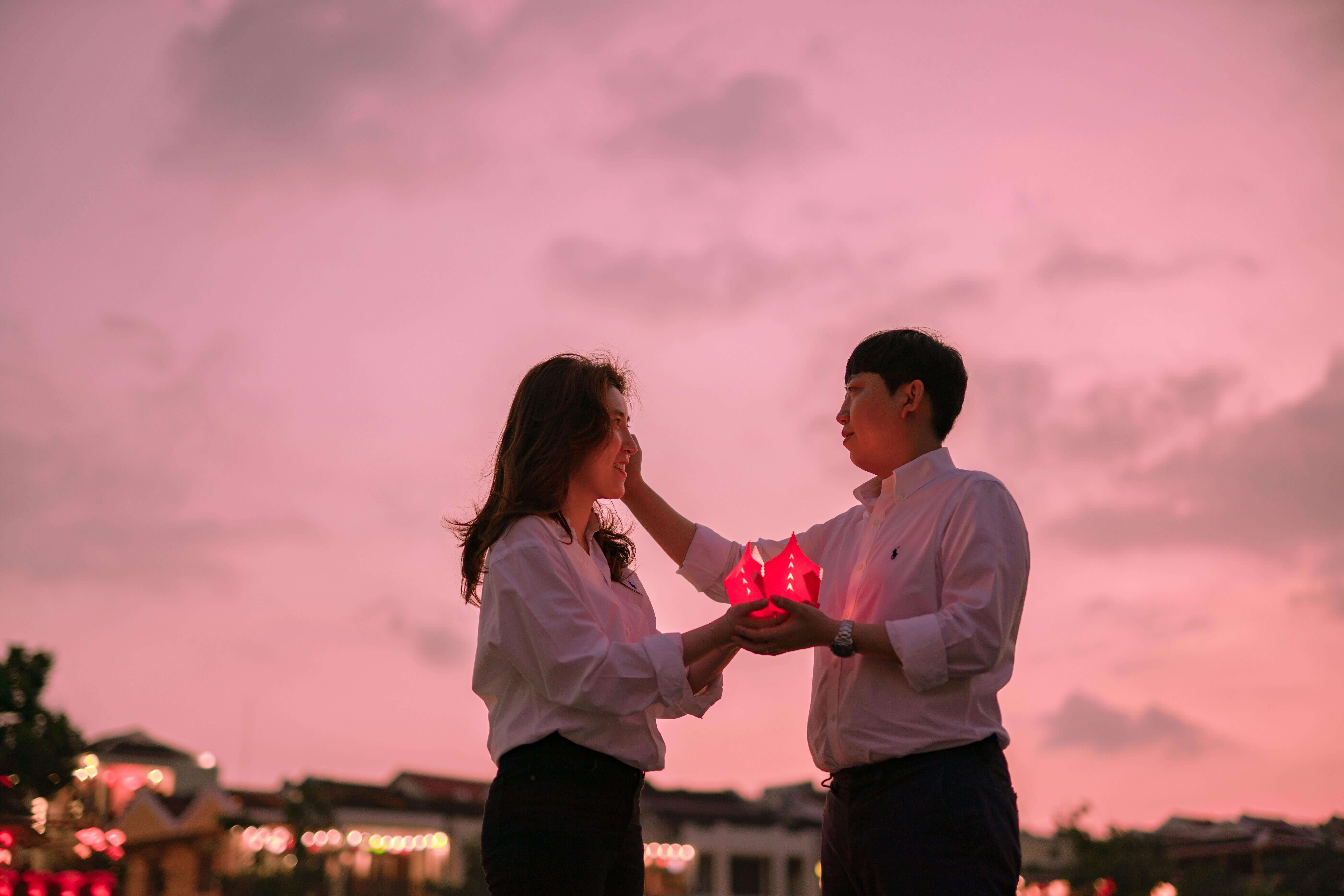 A couple shares a tender moment while holding lanterns against a vibrant dusk sky.