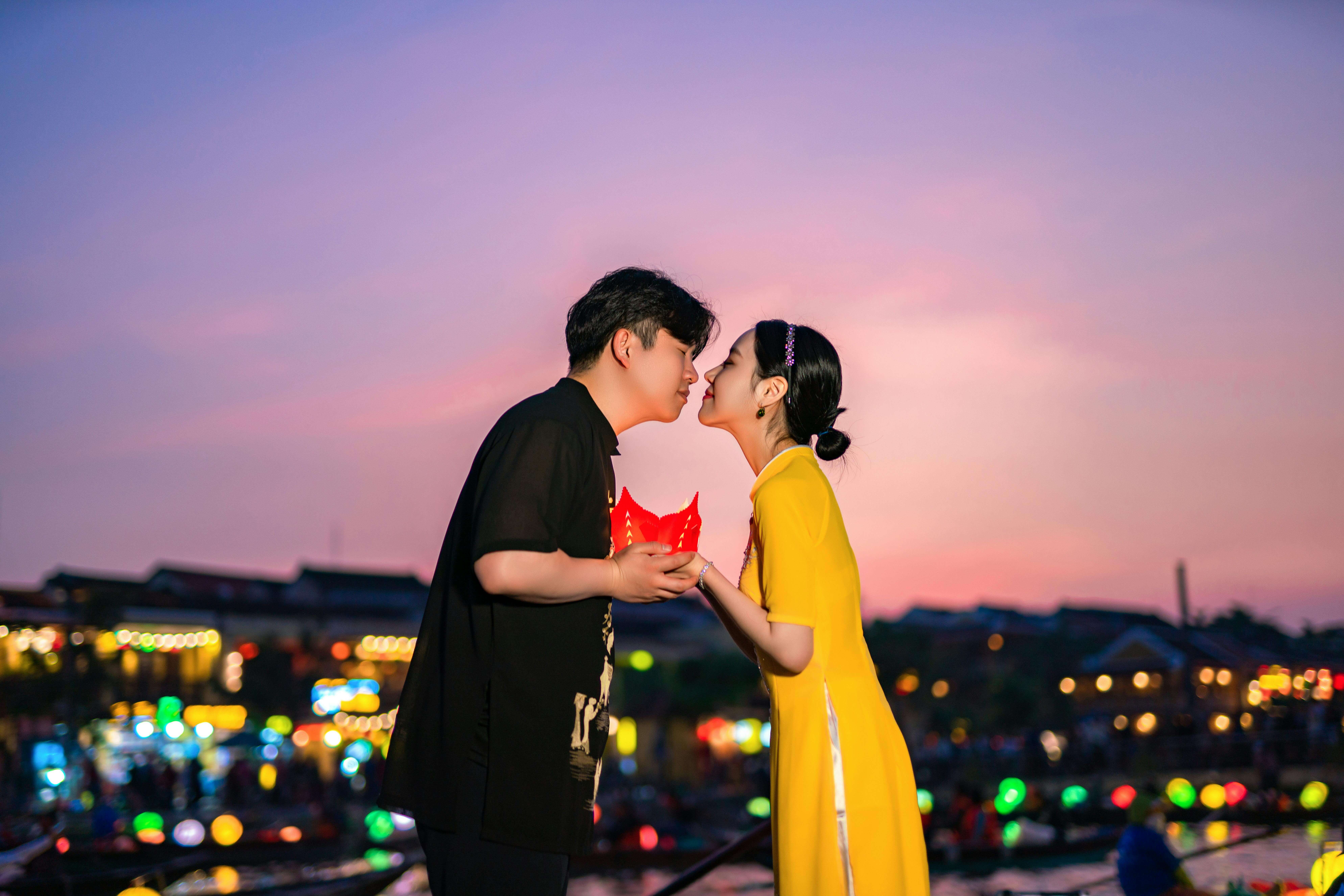 A couple gently kisses, holding a red lantern under a colorful sunset by a vibrant waterfront.