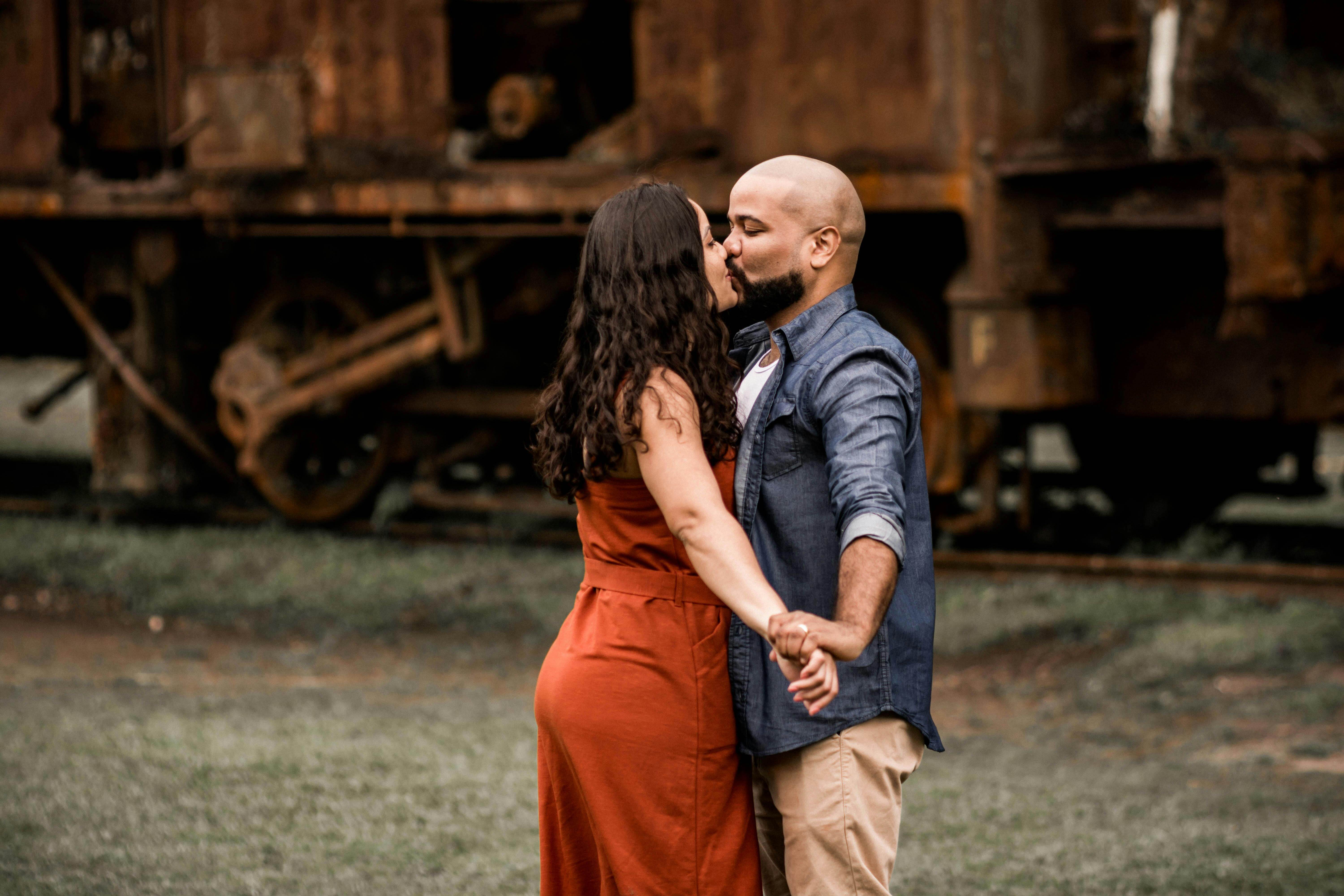 A couple shares a tender kiss in front of an old rusty train, showcasing a moment of affection.
