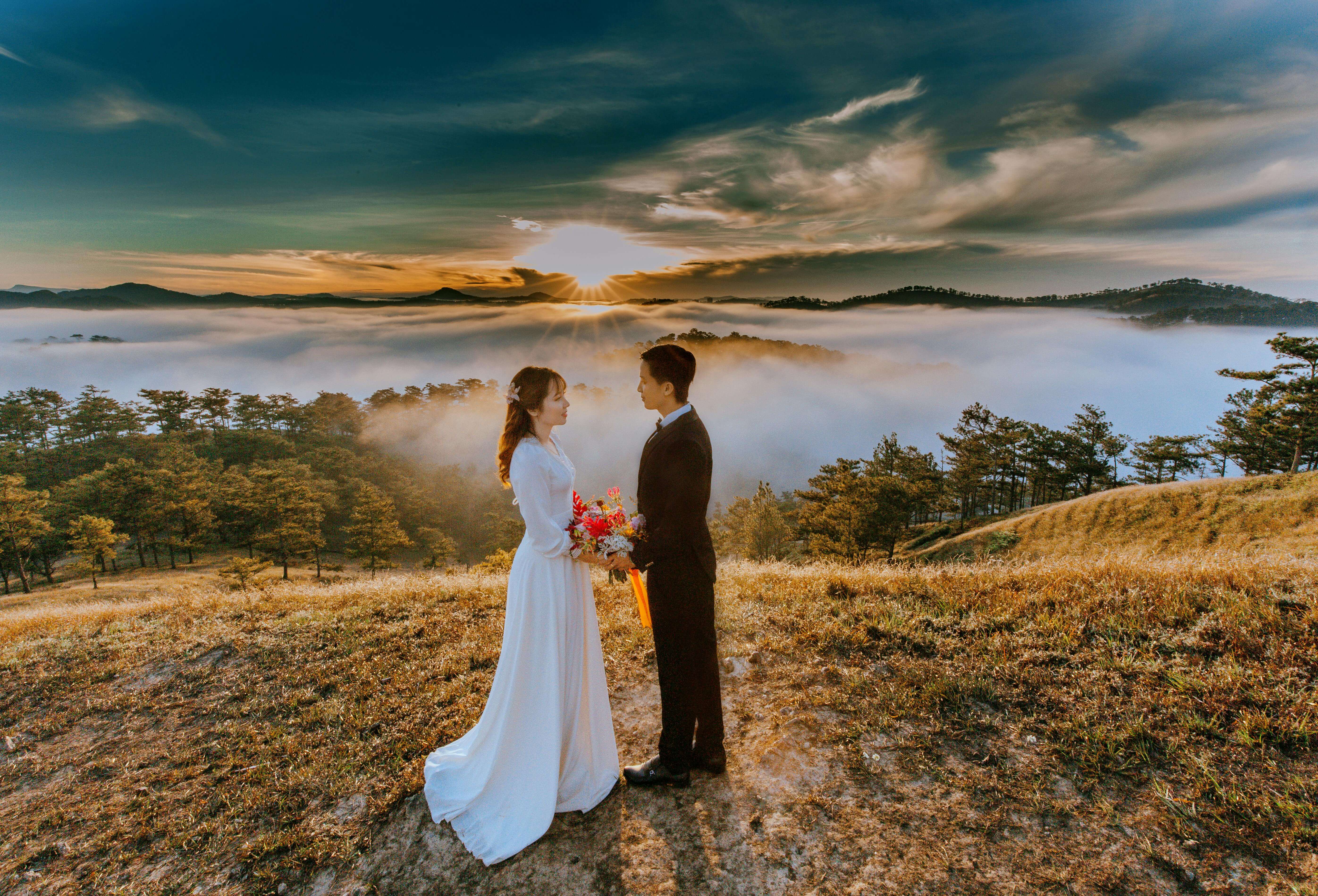 Bride and groom in wedding attire standing in a sea of clouds during sunset, exuding romance and elegance.