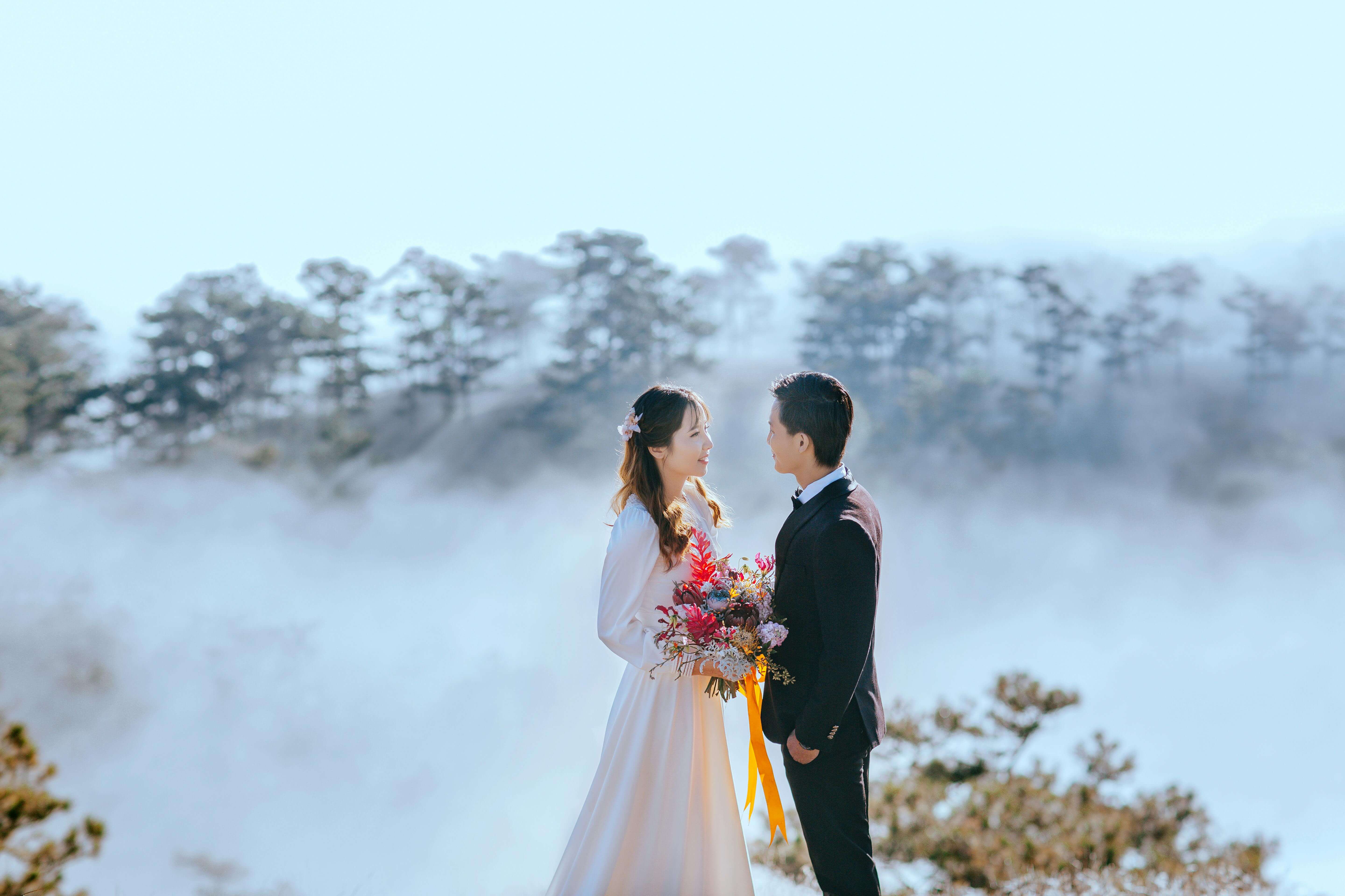 A beautiful Asian couple in wedding attire poses romantically with a scenic foggy forest backdrop.
