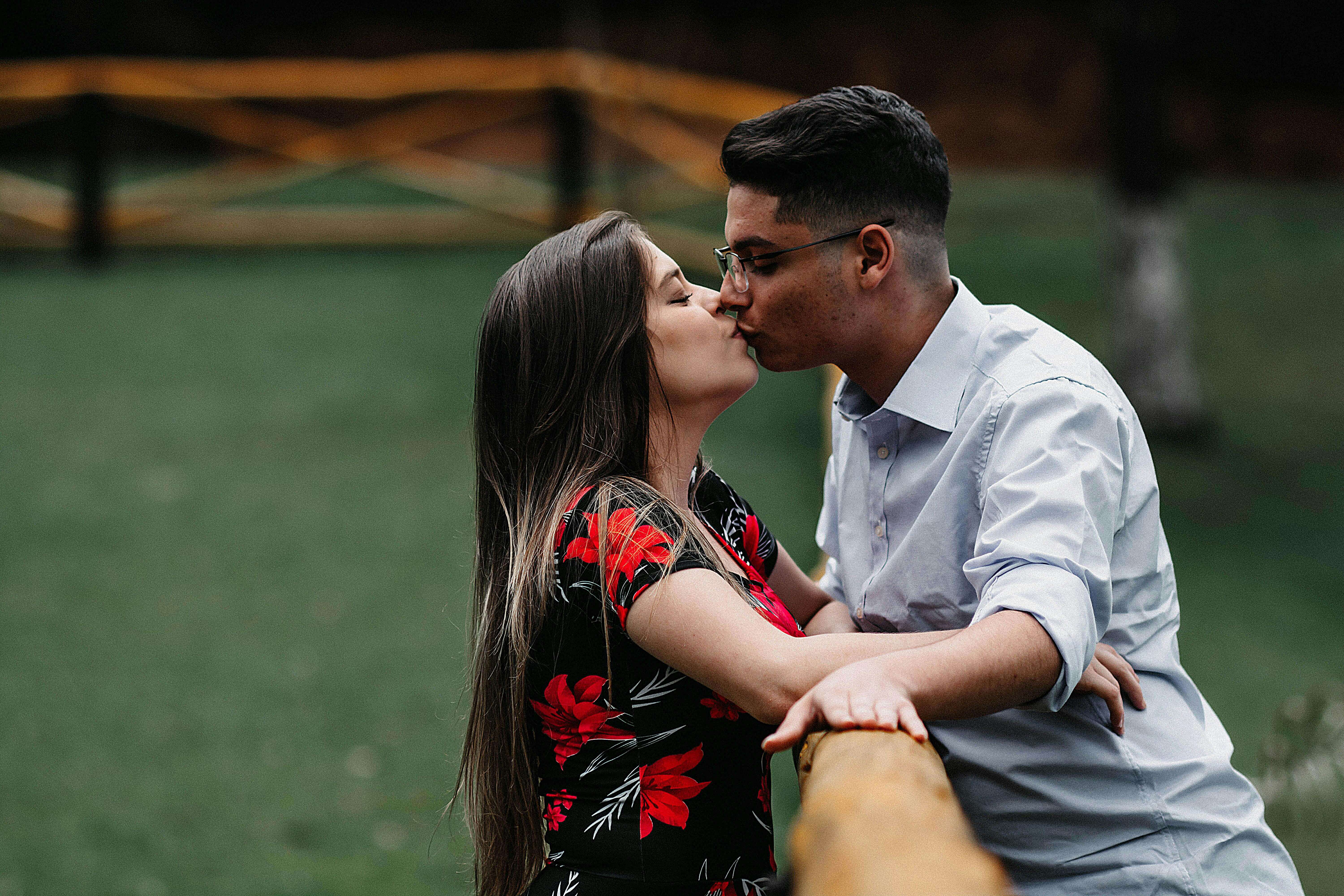 A young couple shares a romantic kiss by a wooden railing in a serene outdoor setting.
