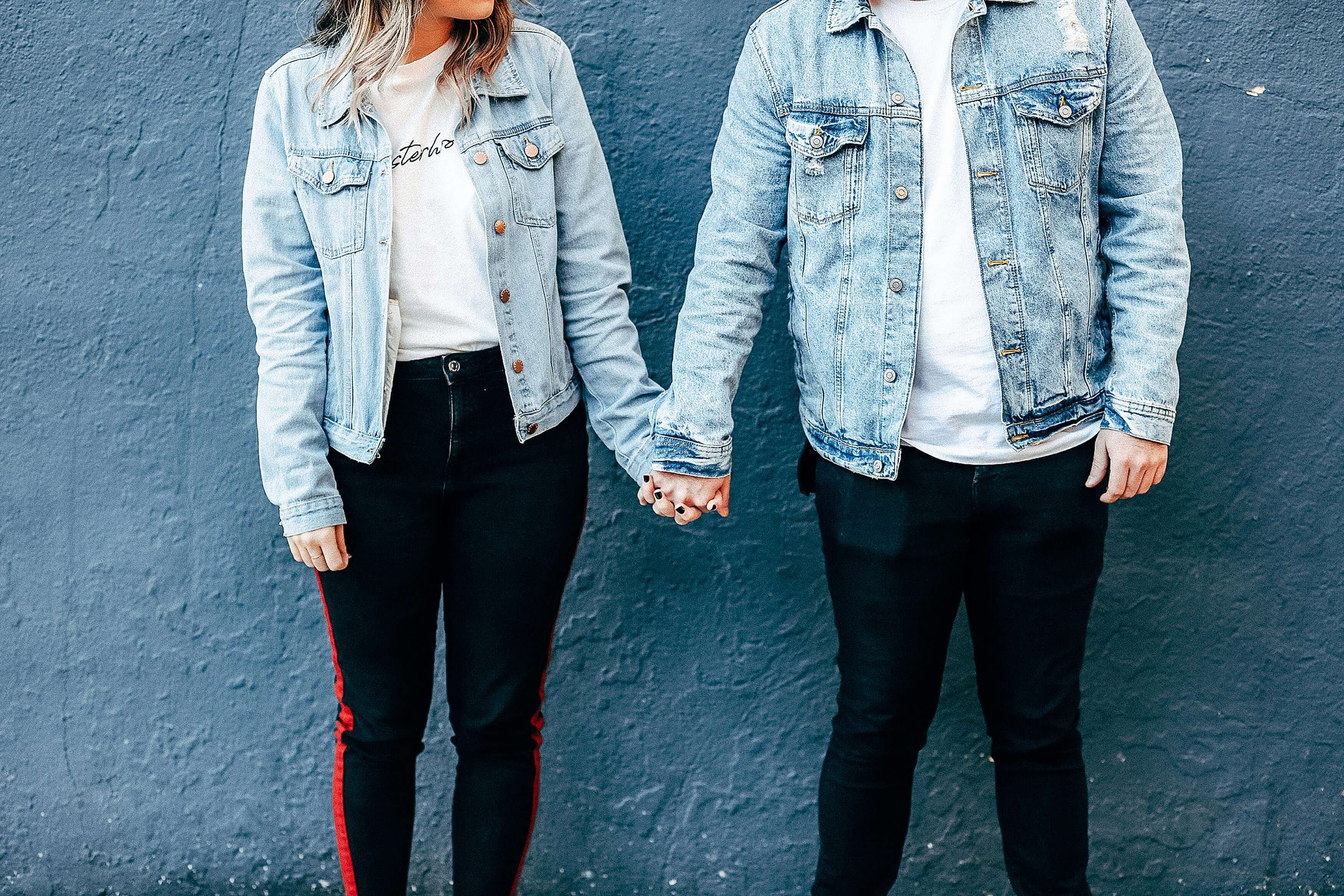 A fashionable couple holds hands against a blue wall, showcasing denim jackets and modern style.