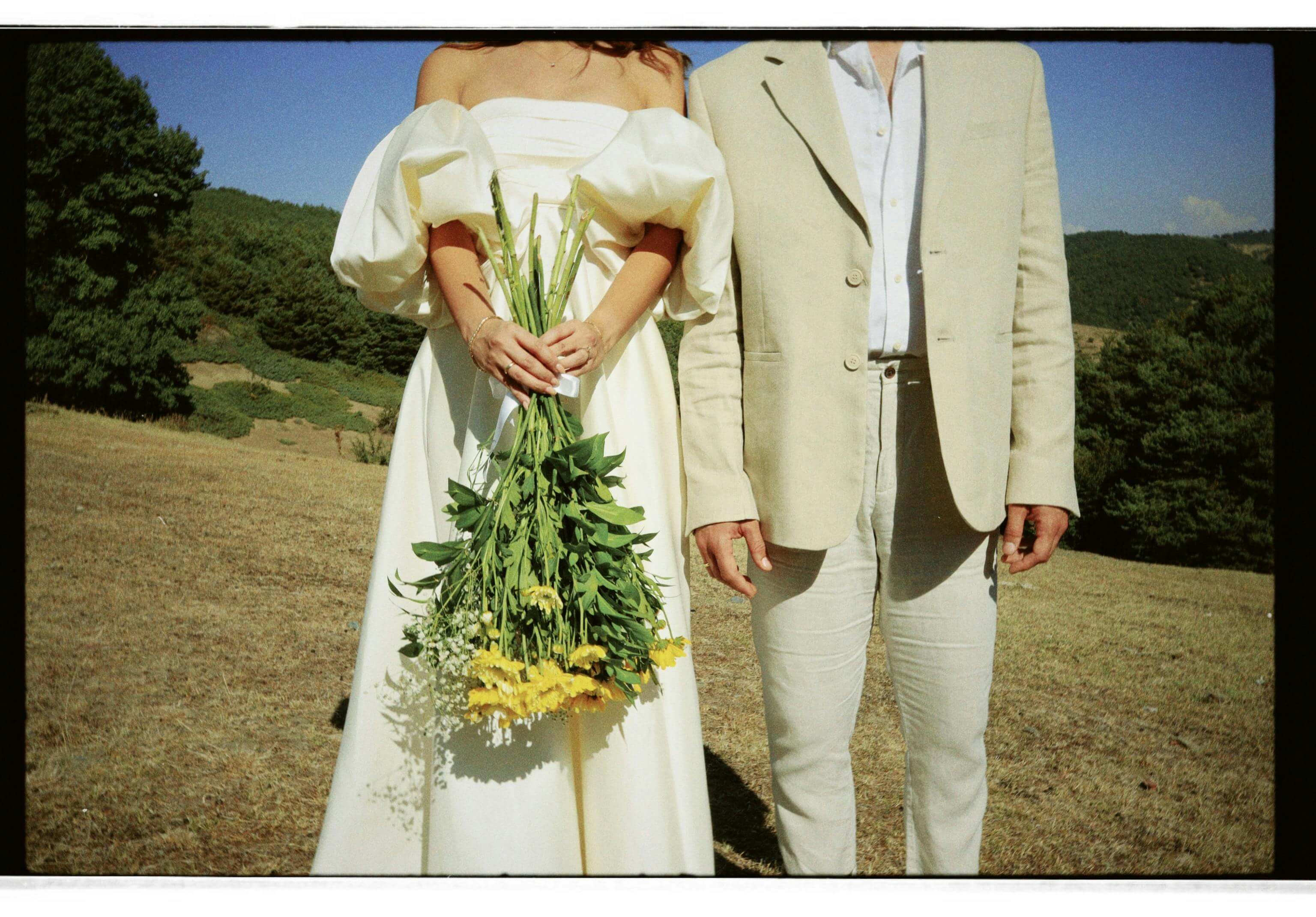A couple dressed in wedding attire stands in an outdoor setting, holding a bouquet.