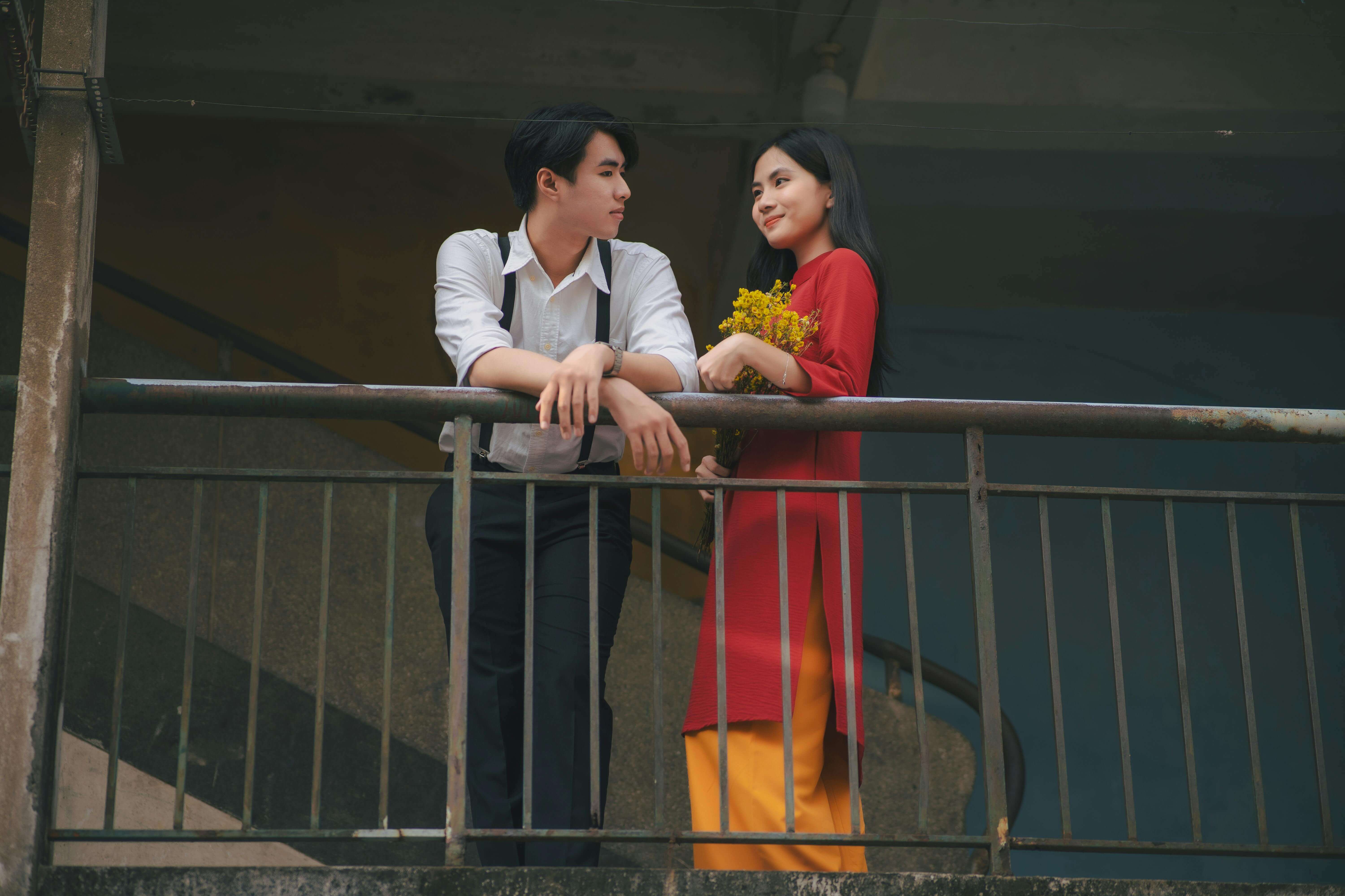 A young couple stands on a balcony sharing a romantic moment with flowers.