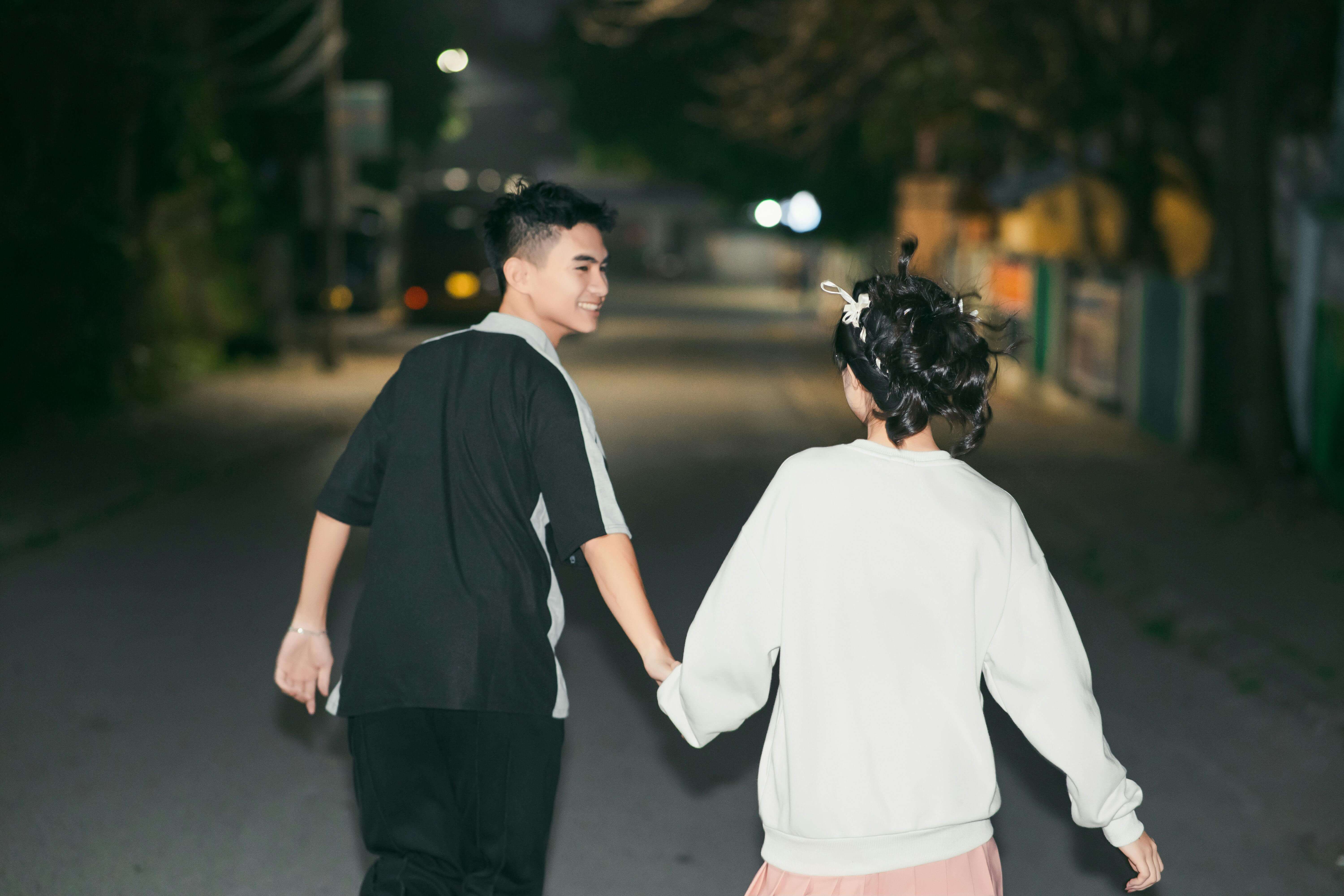 A young couple walks hand-in-hand on a dimly lit street at night, embracing a romantic moment.