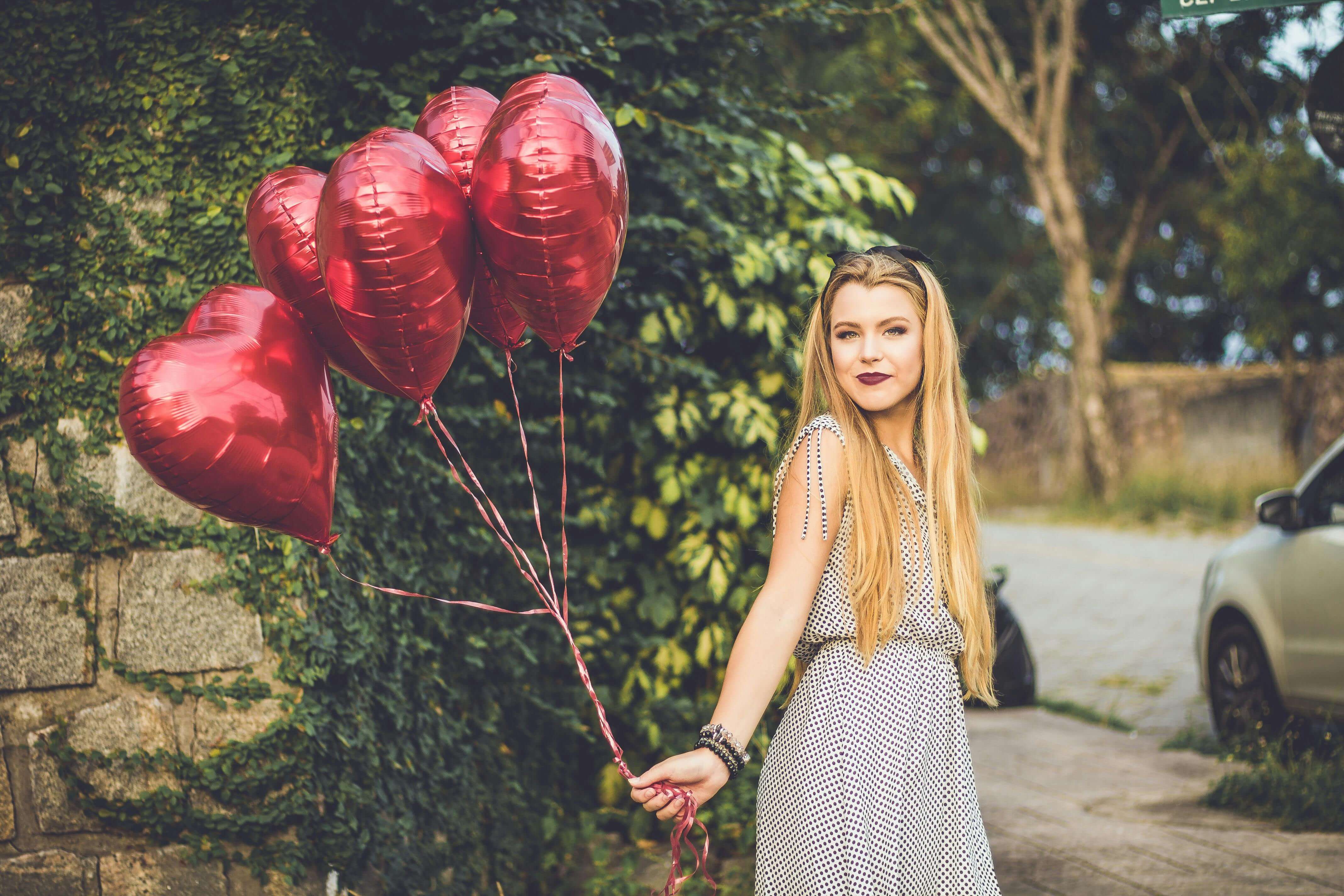 Smiling woman holding red heart balloons in a sunny outdoor setting.