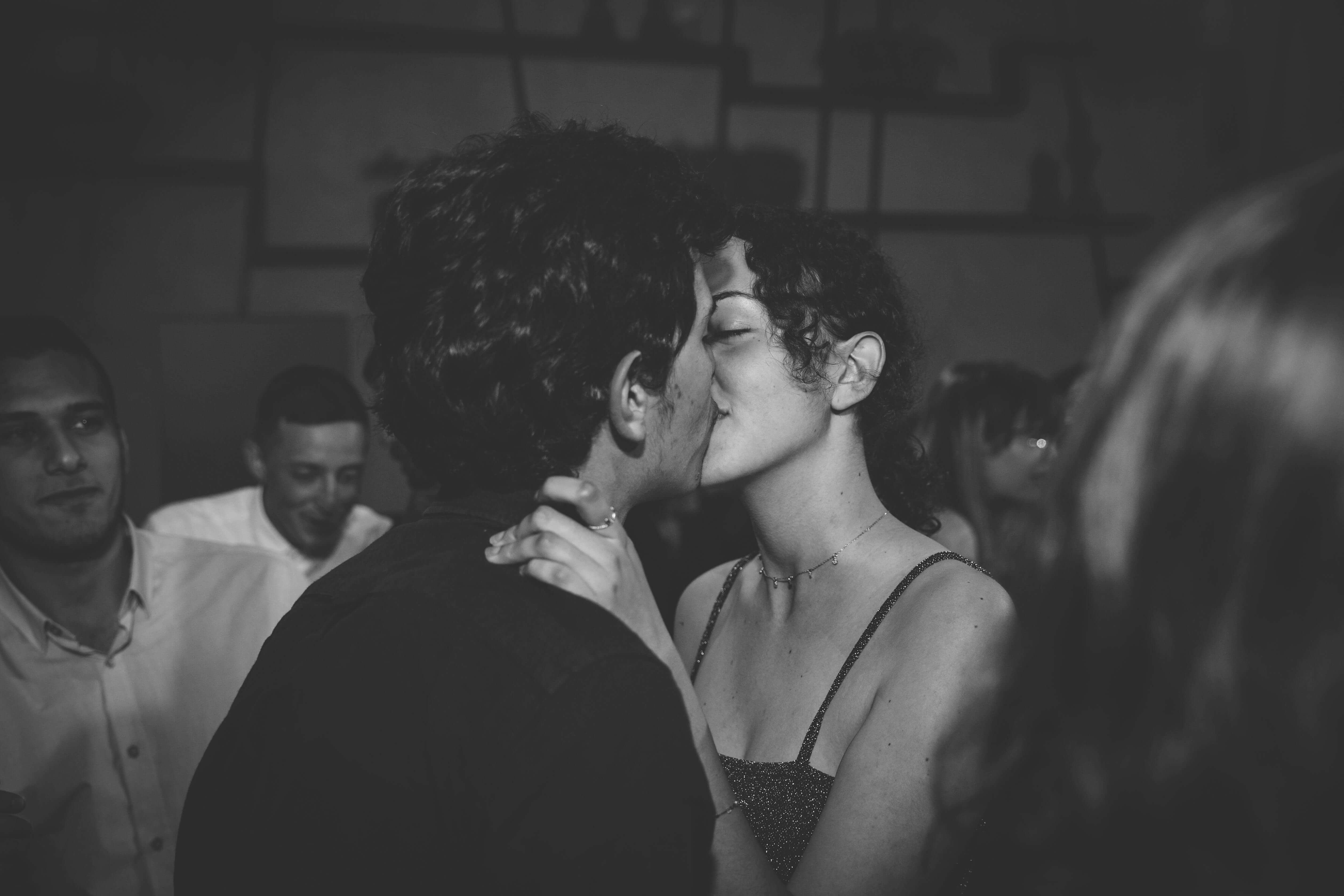 A romantic couple shares a kiss at a lively indoor party, captured in black and white.