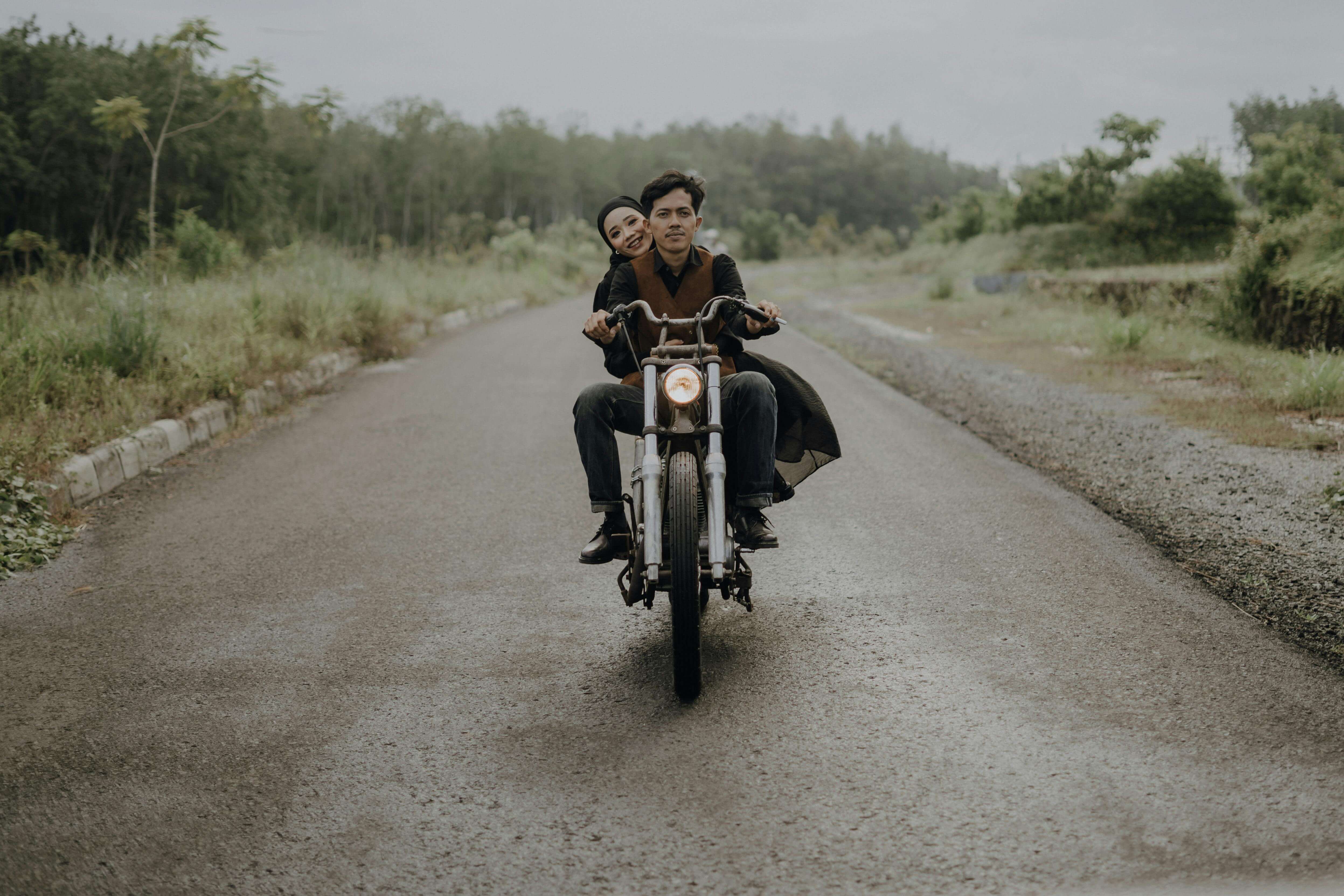 Asian couple riding a motorbike on a rural road amidst lush greenery in Jakarta, Indonesia.
