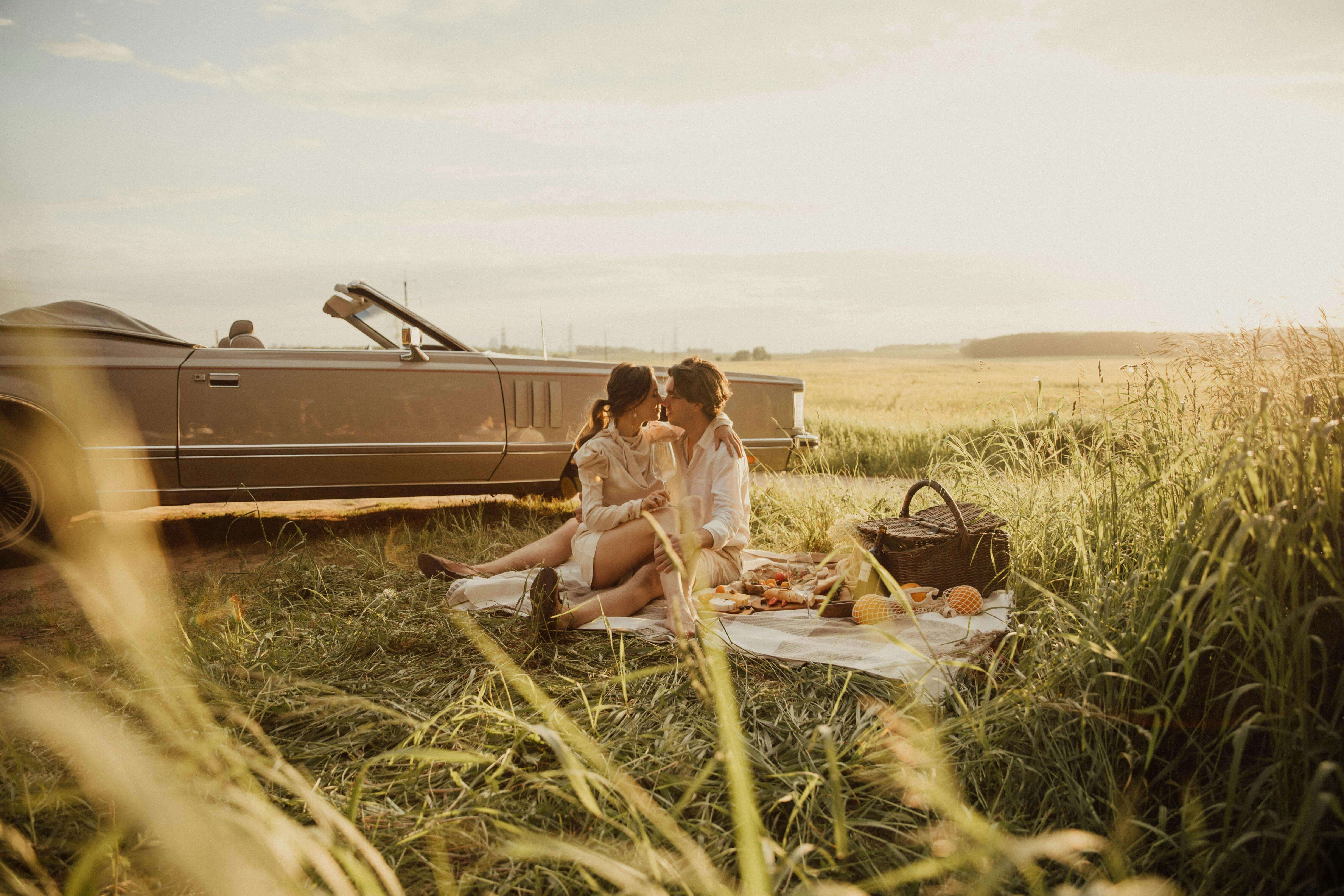 Young couple enjoying a romantic picnic beside a vintage convertible in a sunny meadow.