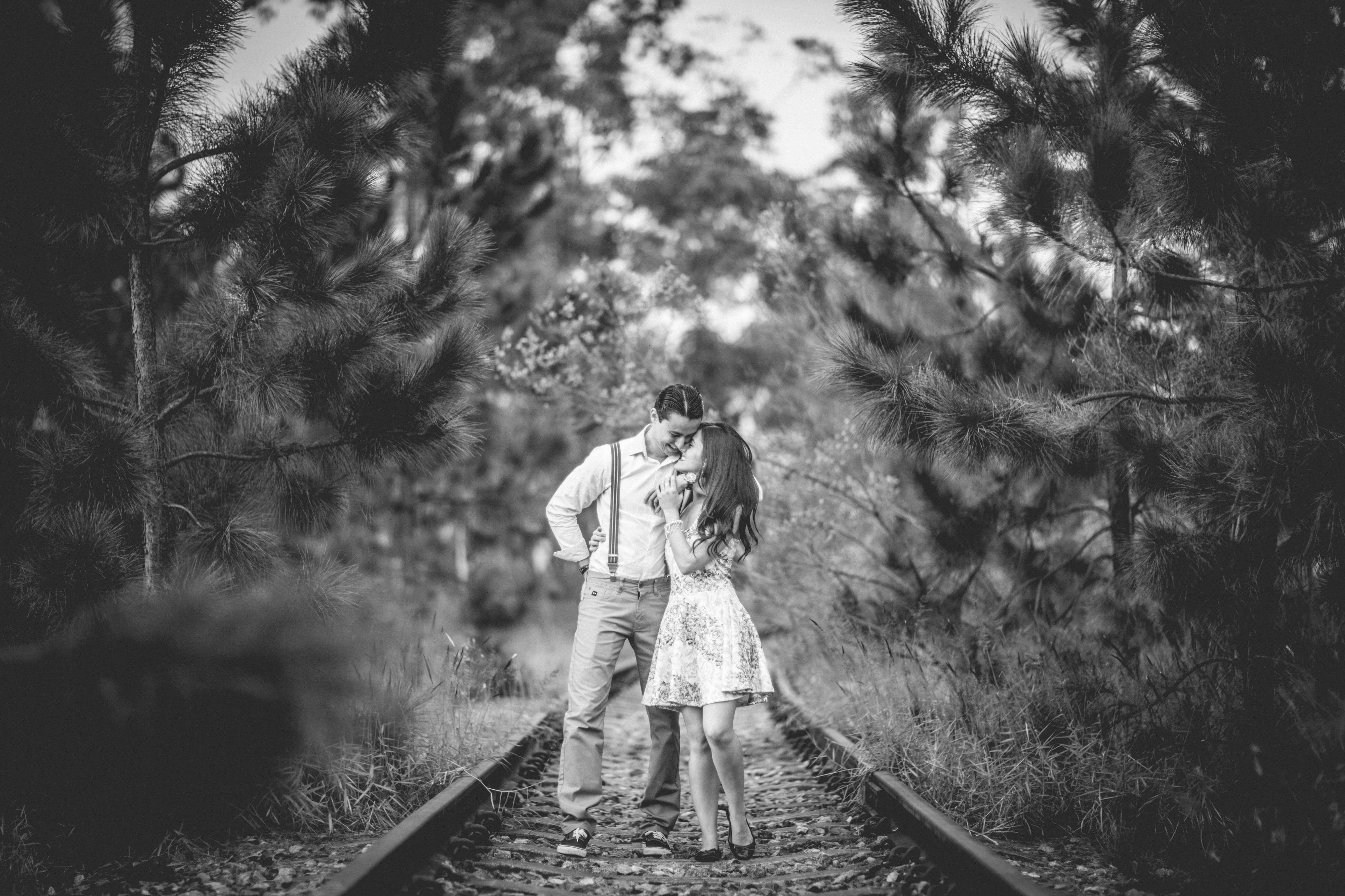 A romantic black and white photo of a couple kissing on railway tracks surrounded by pine trees.