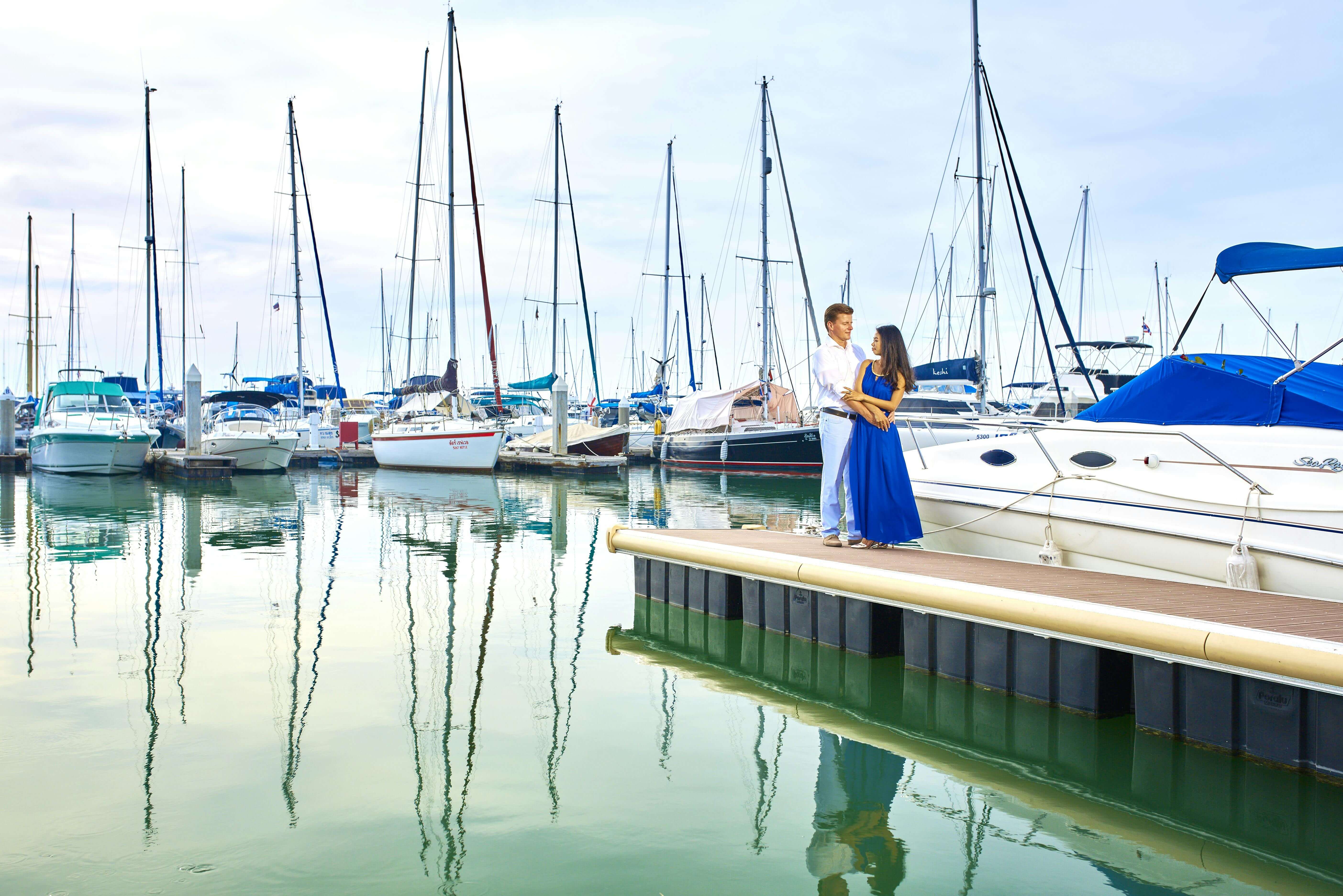 A couple standing on a dock in a marina, surrounded by sailboats, enjoying a romantic moment.