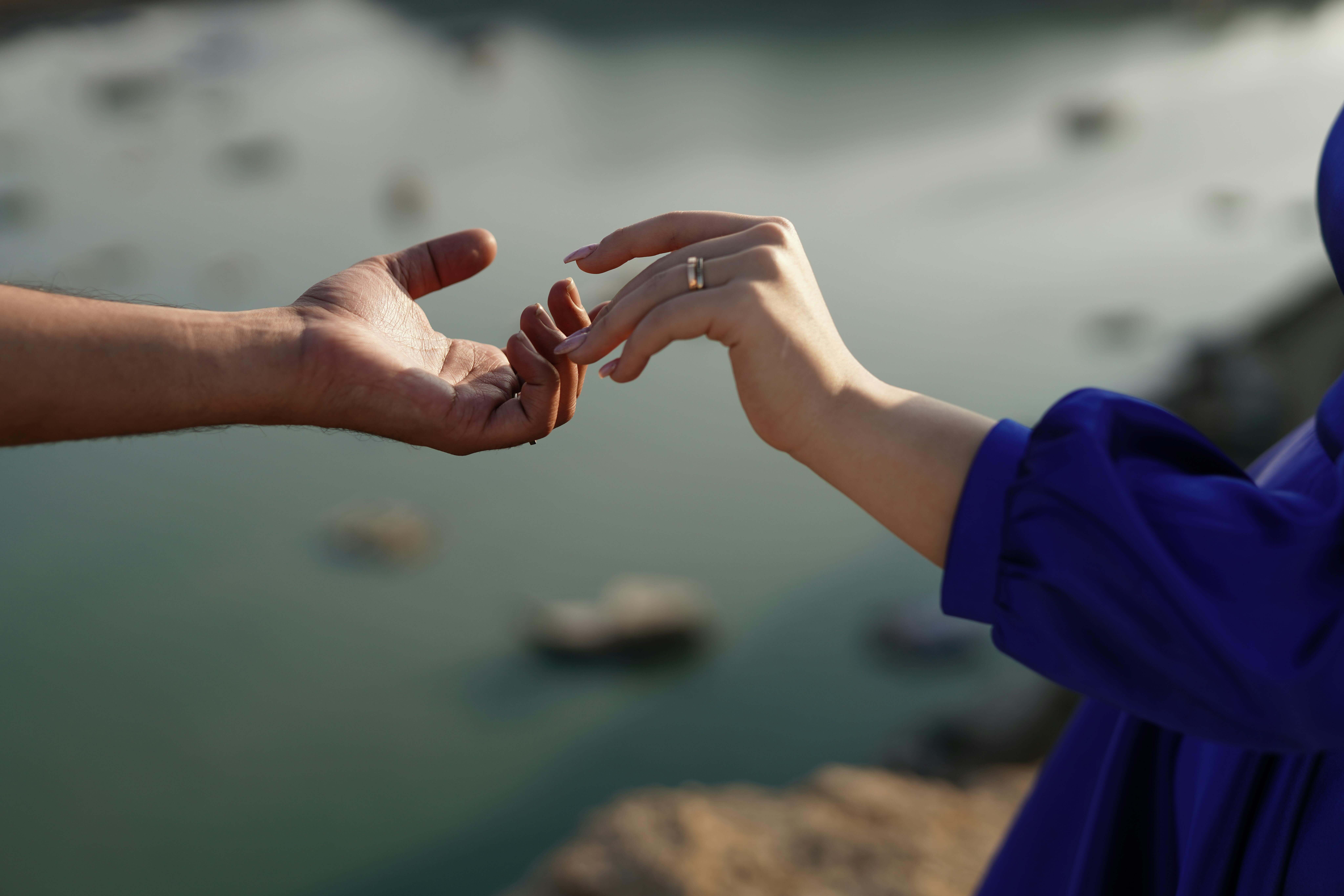 A man's and a woman's hands reaching towards each other, set against a serene water landscape.