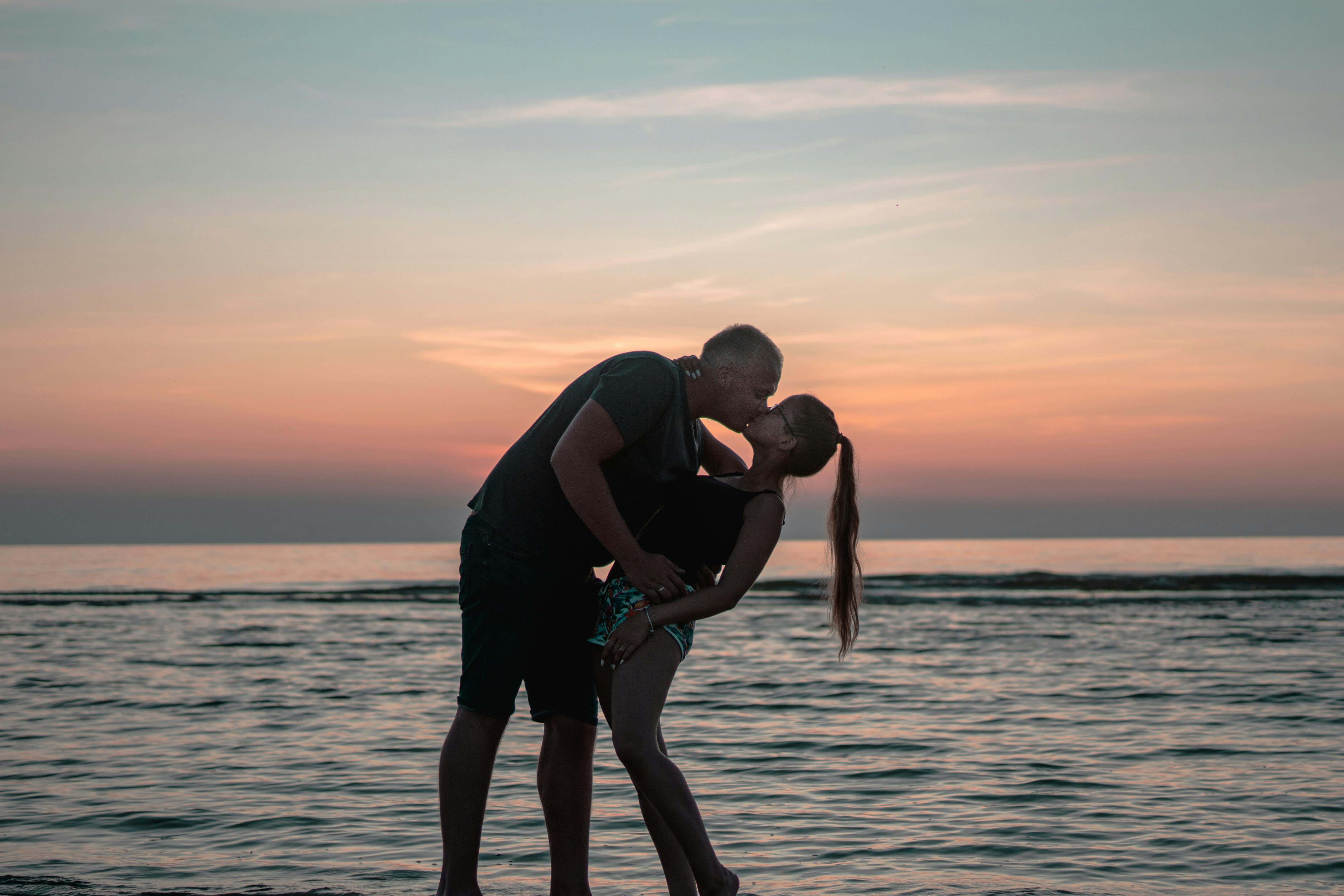 A couple shares a romantic kiss on the beach during a vibrant sunset by the ocean.