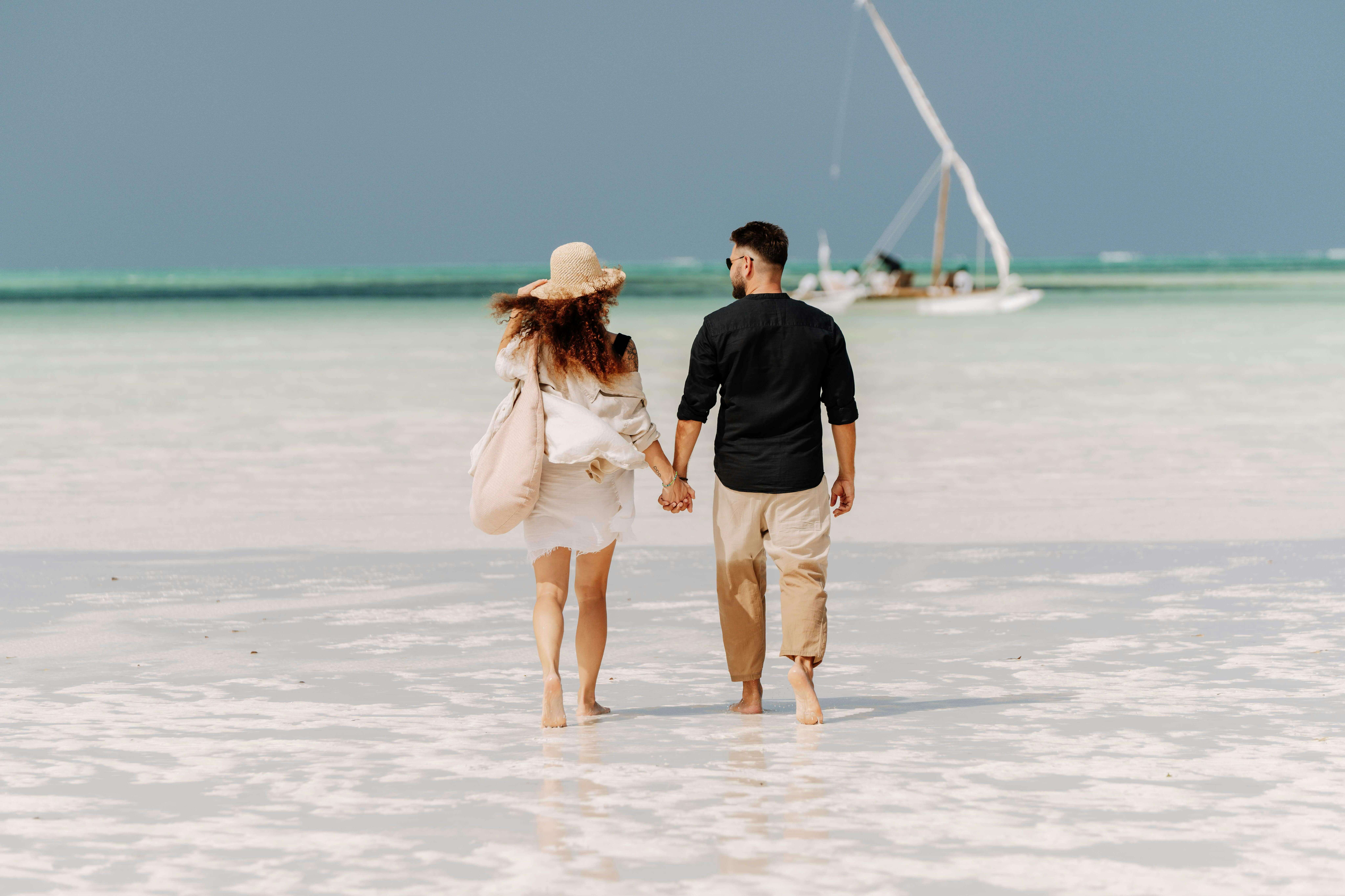 Couple walking hand-in-hand on a sunny Zanzibar beach, with a traditional dhow in the background.