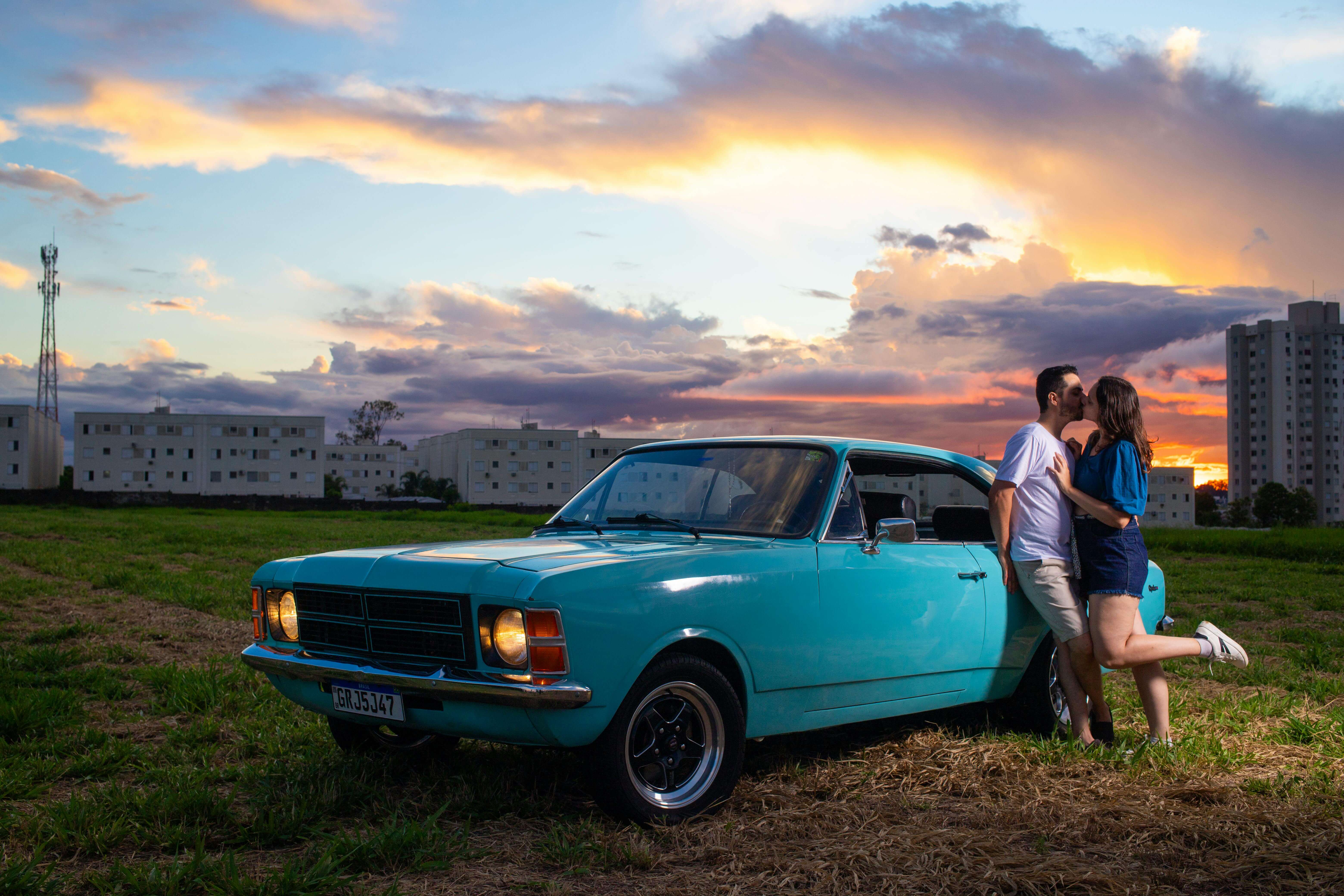 A couple shares a romantic moment beside a vintage blue car during a scenic sunset in a city field.