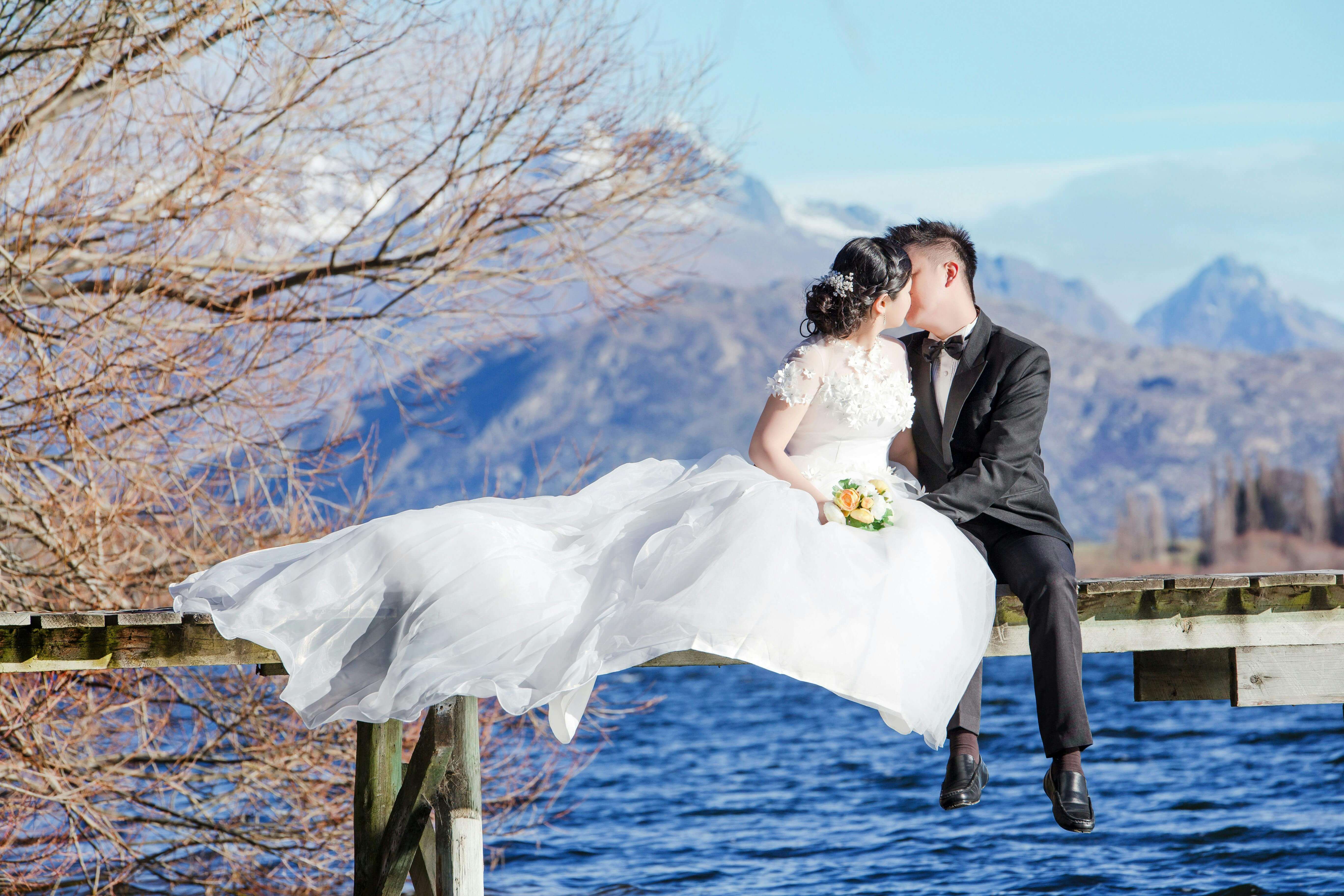 Beautiful wedding couple in love kissing on a dock by the lake against a stunning mountain backdrop.
