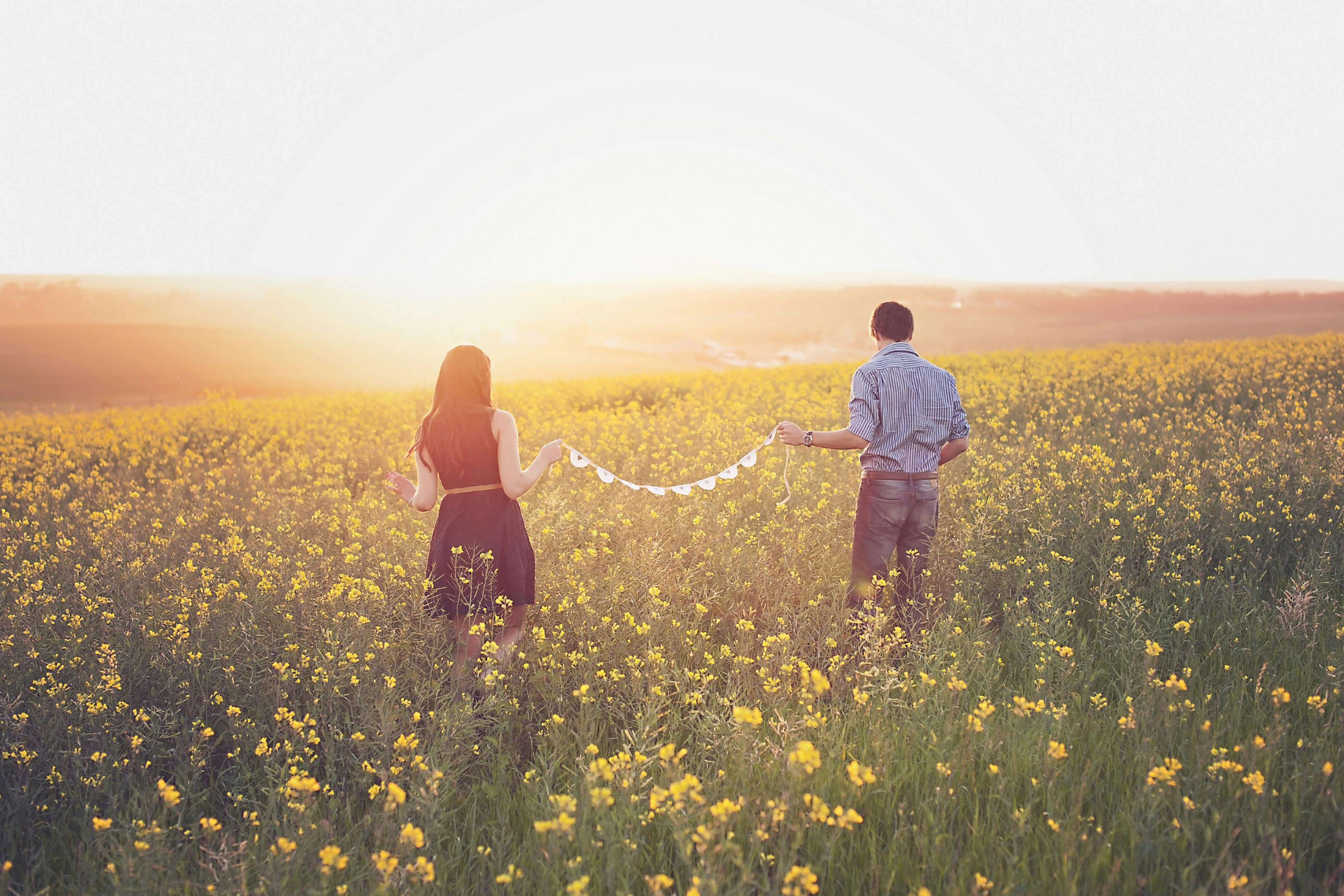 A couple enjoys a romantic walk through a sunlit flower field at sunset, holding a string of hearts.