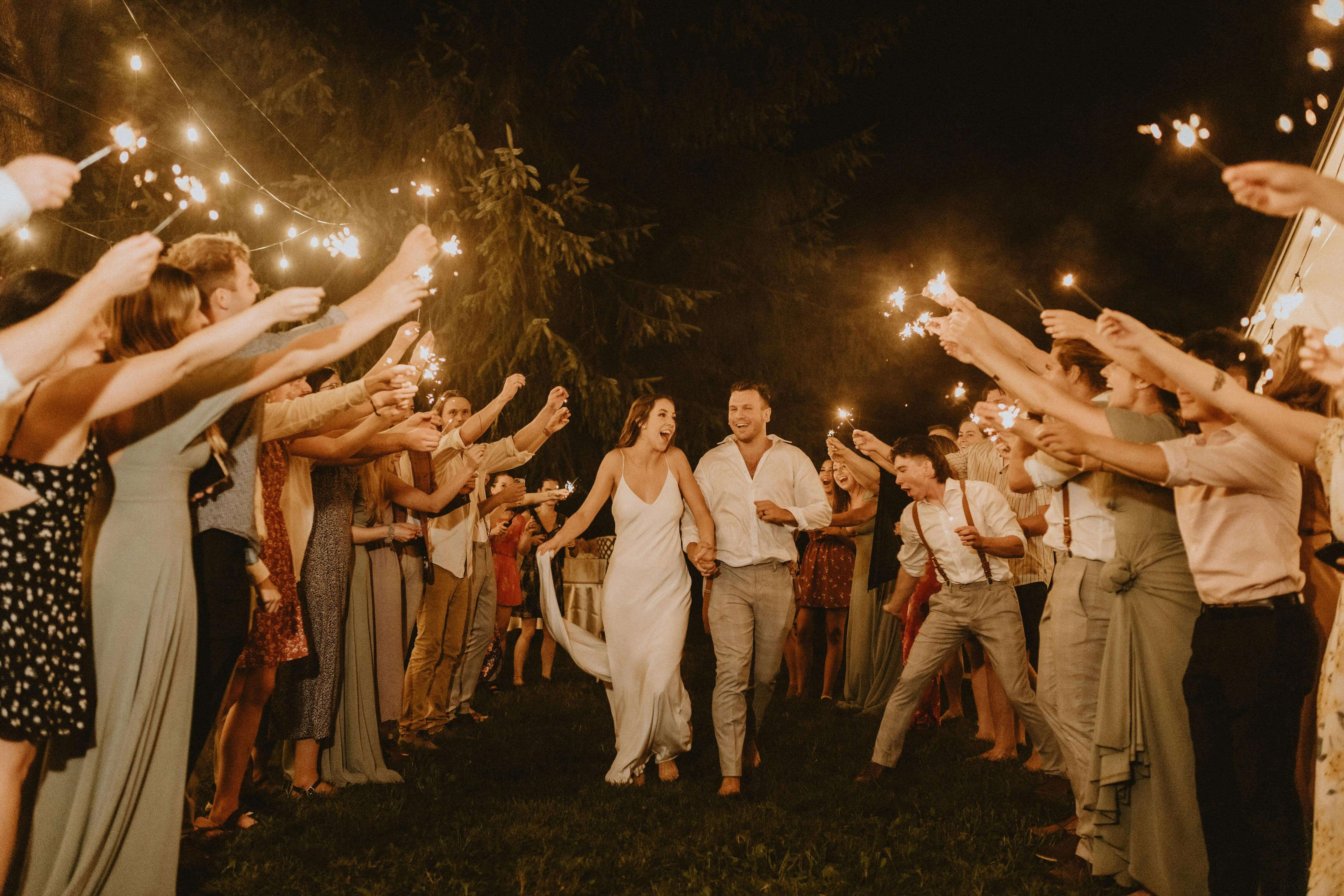 Bride and groom joyfully exit surrounded by sparkling celebratory guests at a nighttime wedding.