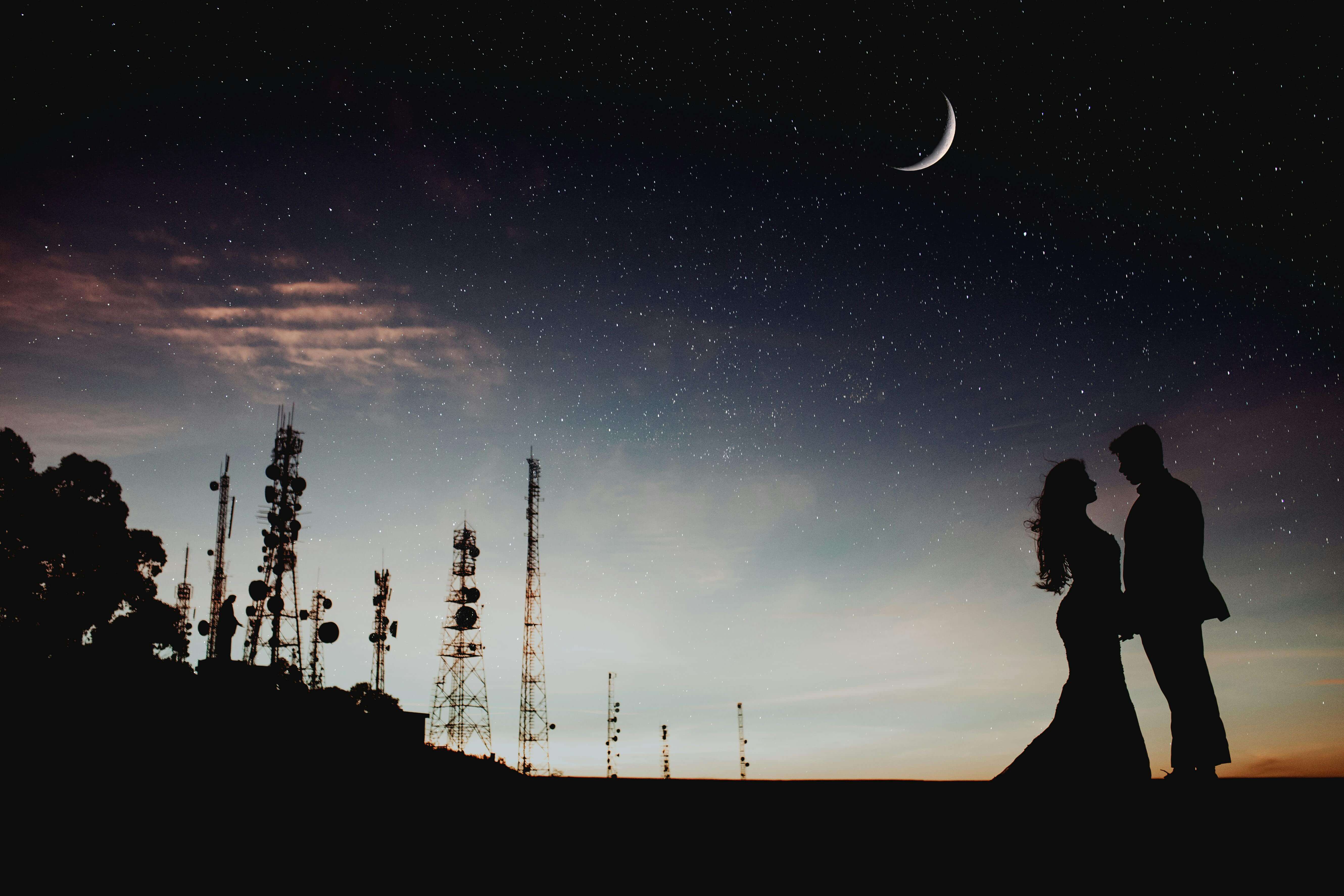 A couple's silhouette against a starry sky with a crescent moon and communication towers, creating a romantic ambiance.