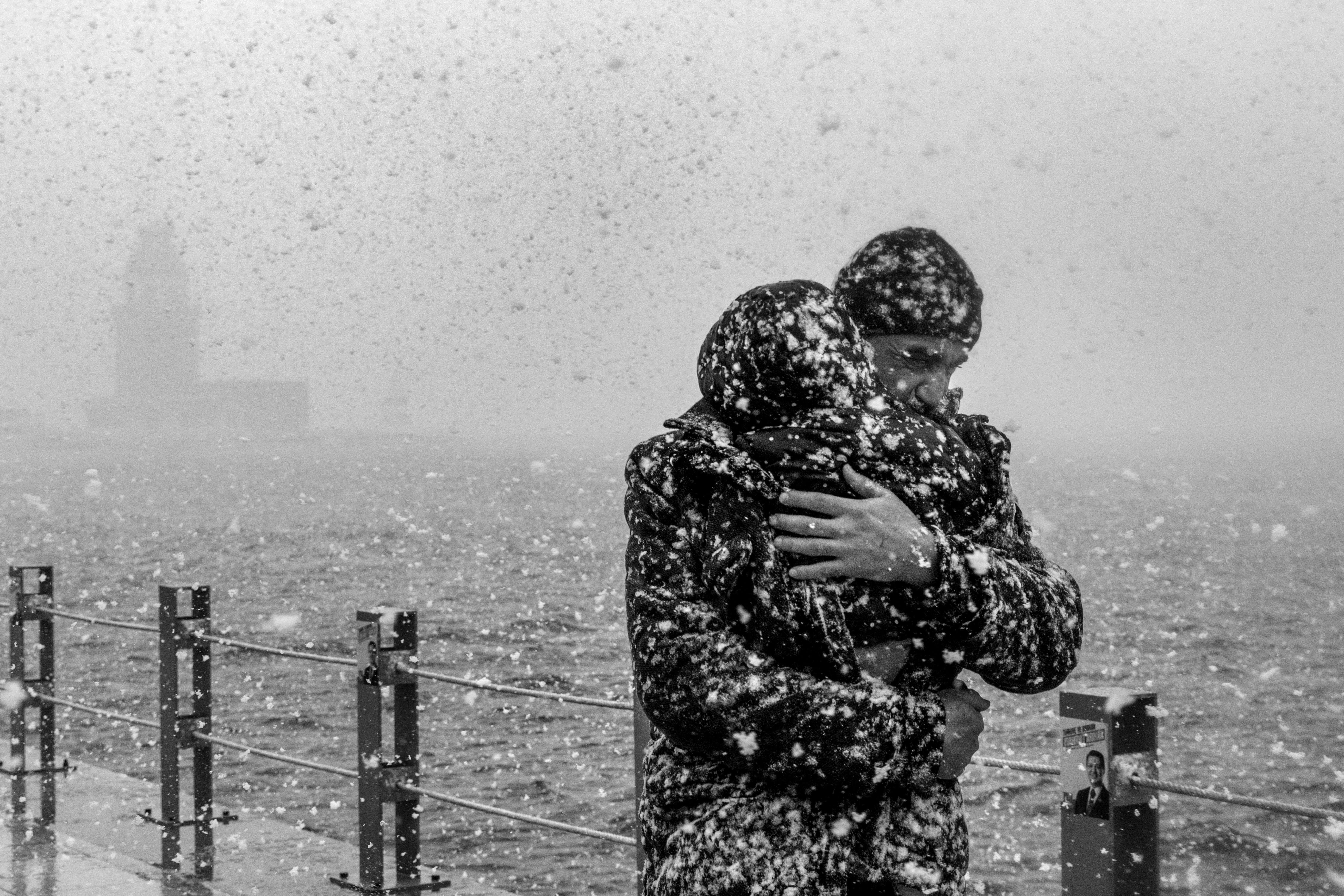 A moving black and white photo of a heartfelt embrace during a snowfall at the waterfront.