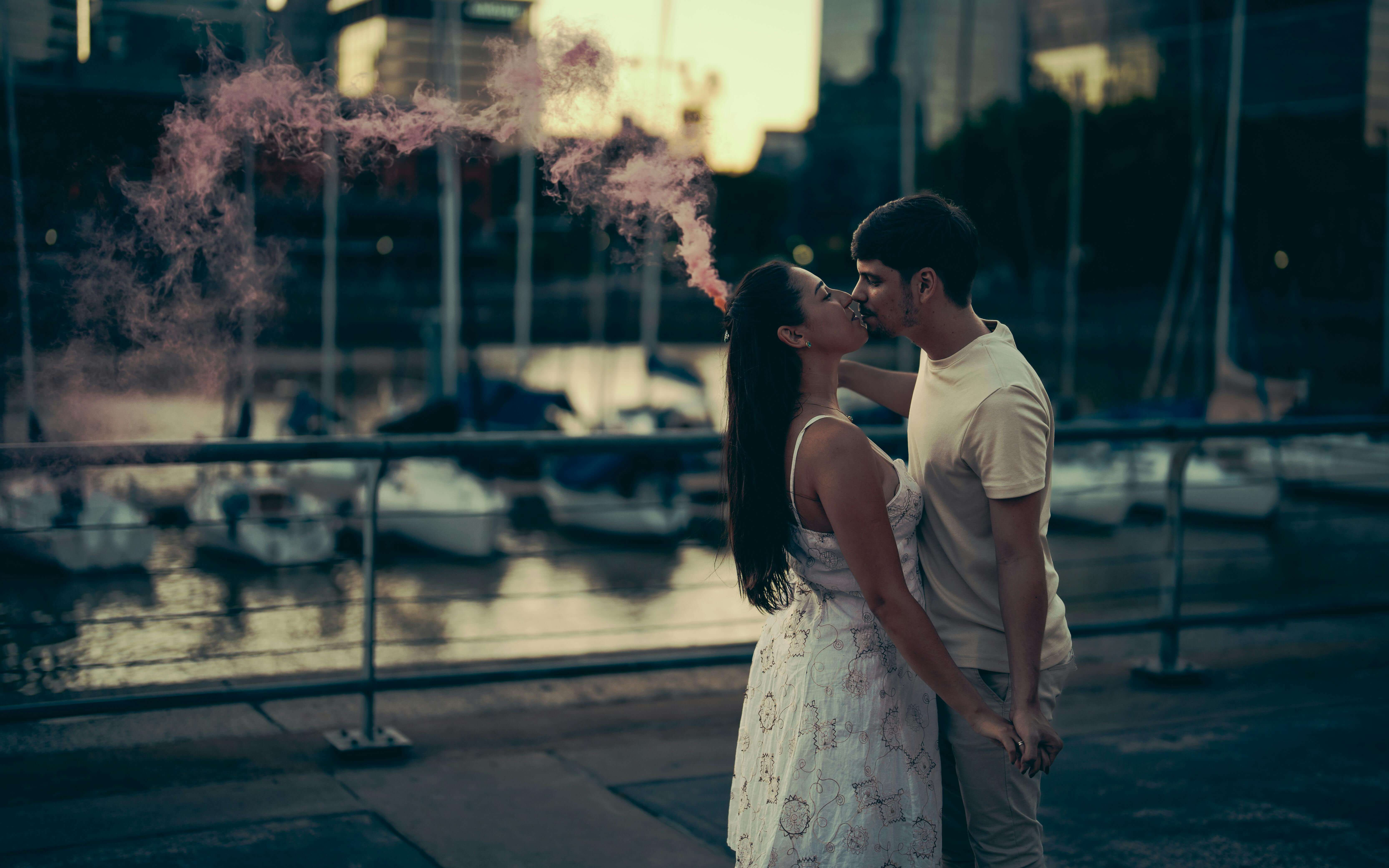 Romantic couple shares a kiss by the marina at sunset with pink smoke in the background.