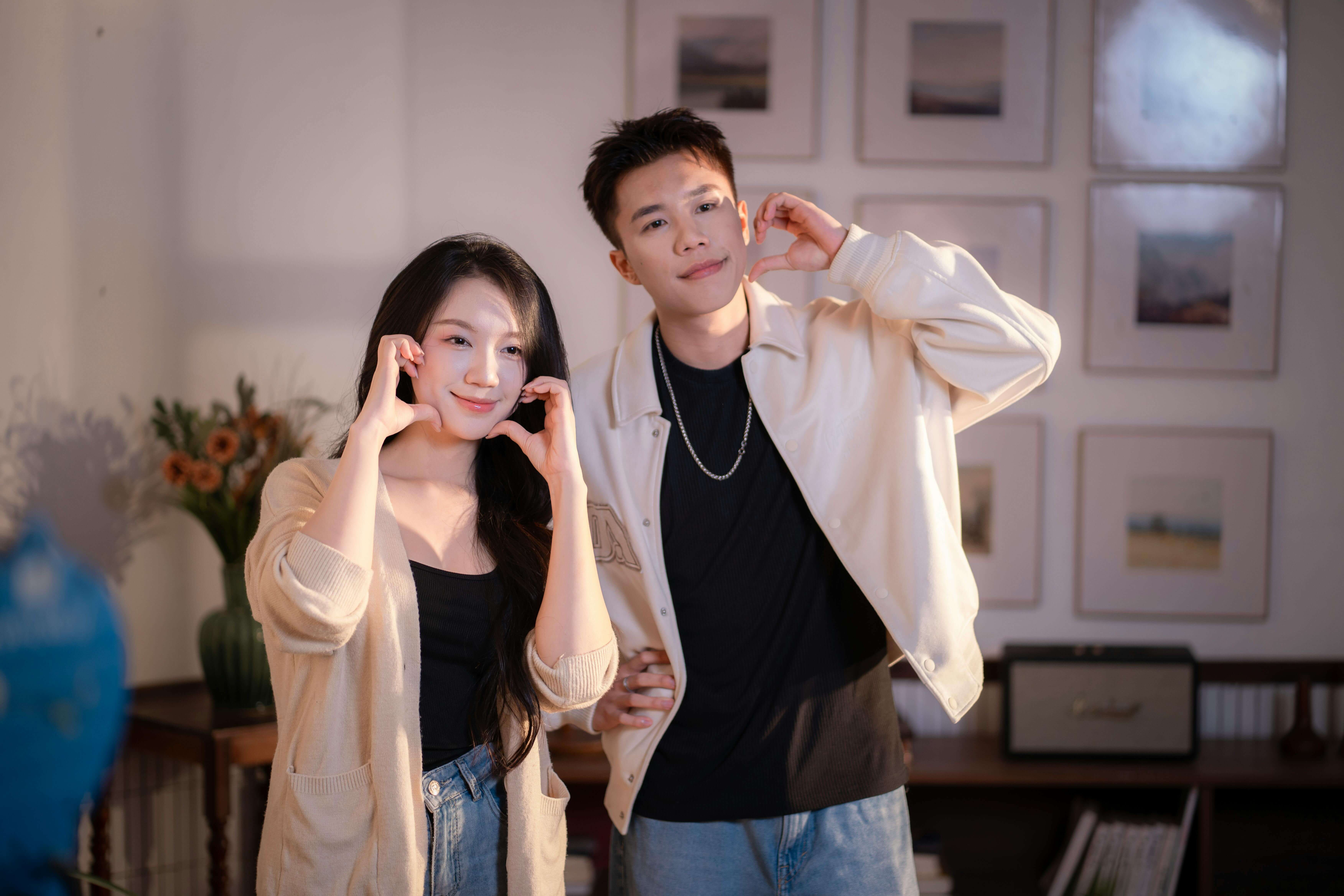 A cheerful couple making heart gestures inside a cozy room with flowers and framed art.