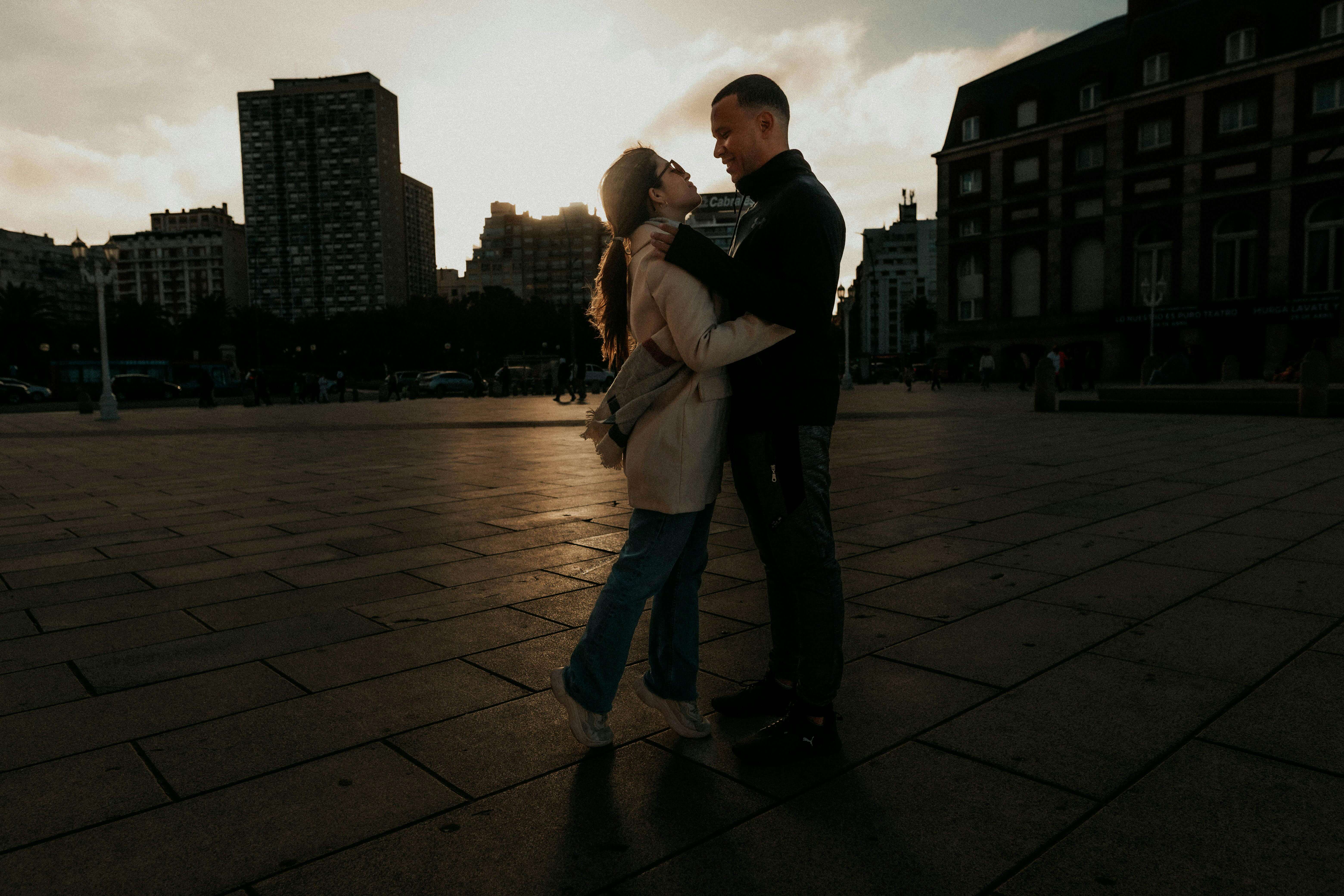A couple embraces in a romantic setting against a sunset backdrop in the city.