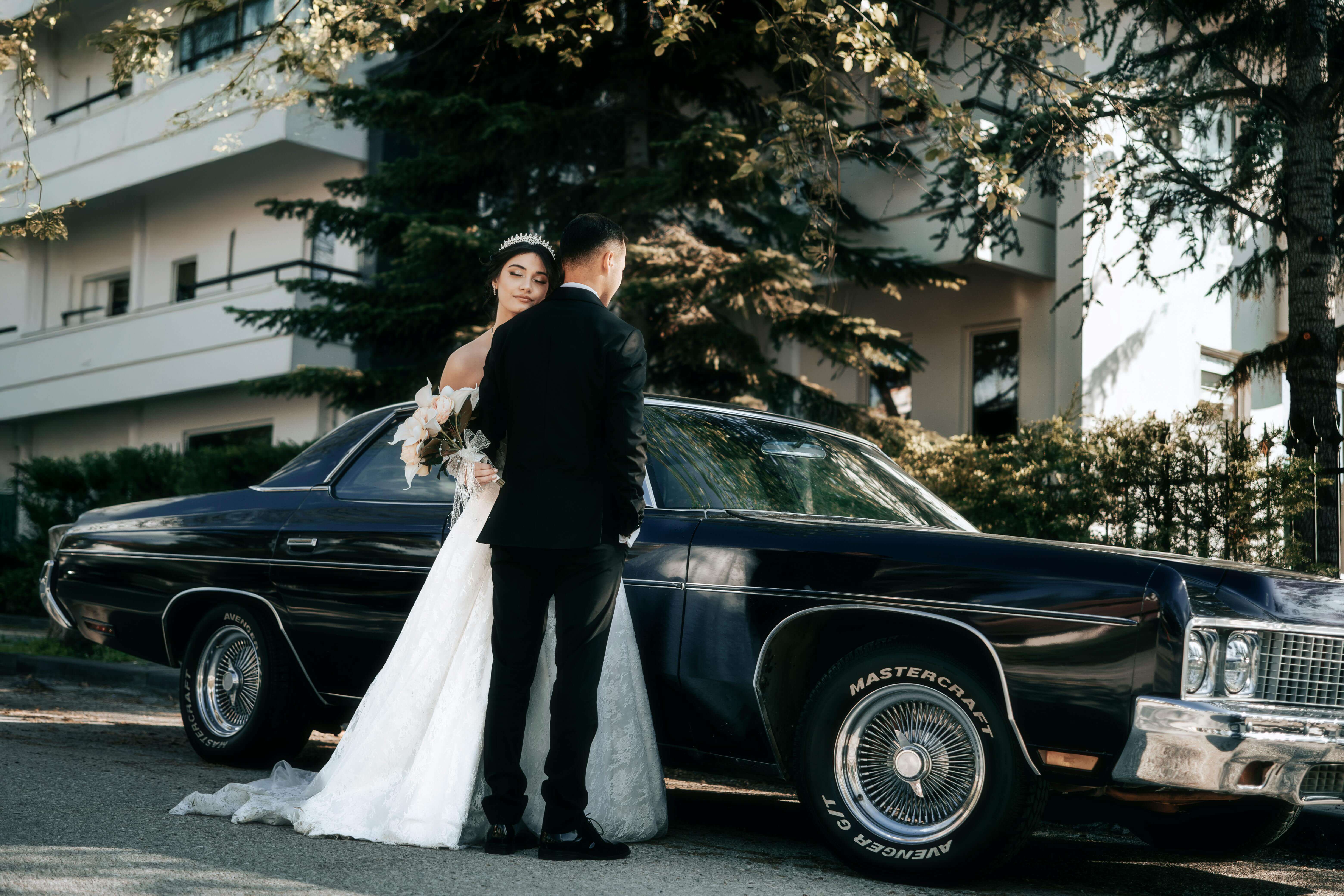 A bride and groom pose by a vintage car outdoors on their wedding day.