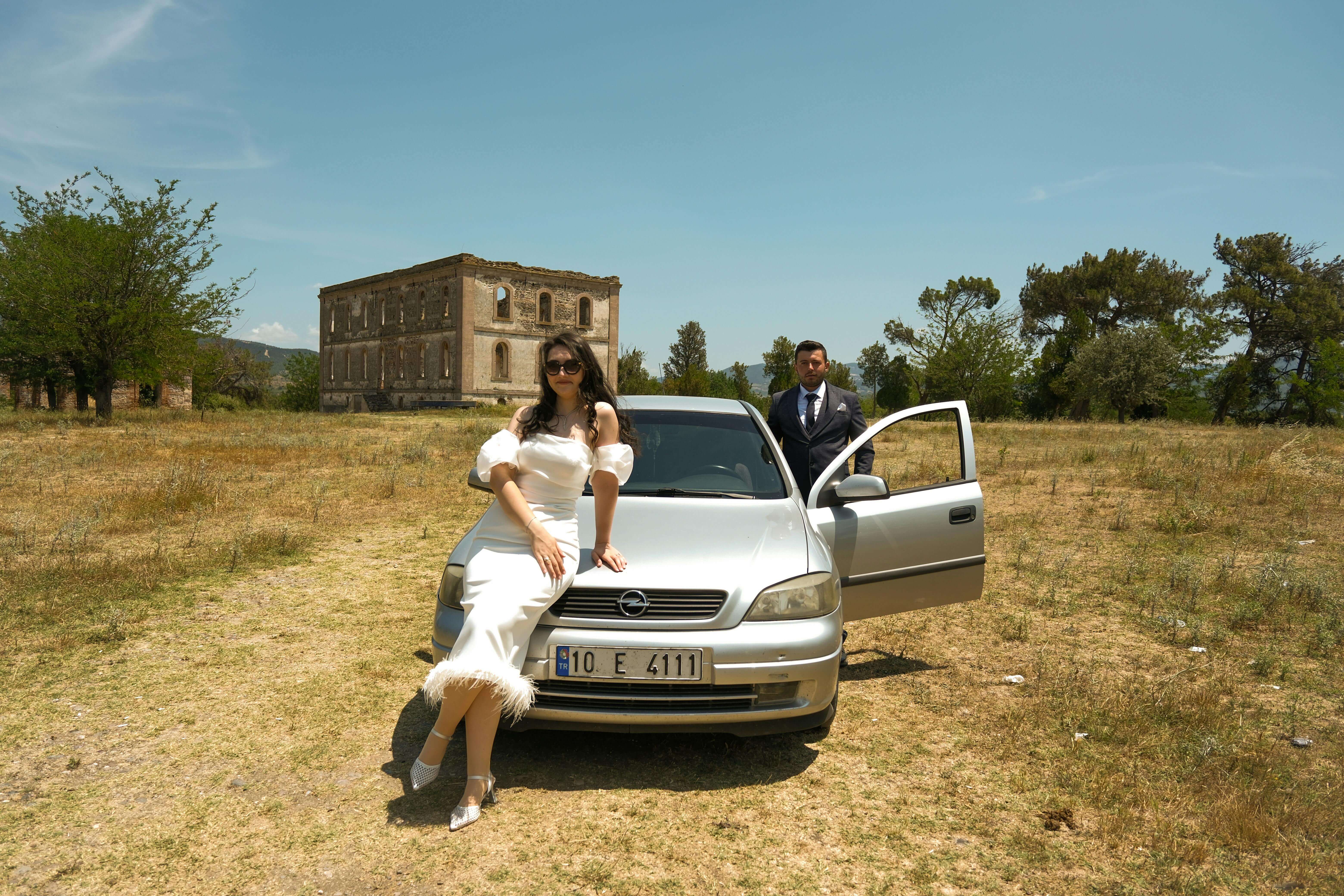 Stylish couple in elegant attire posing with a silver car in front of a historic building.