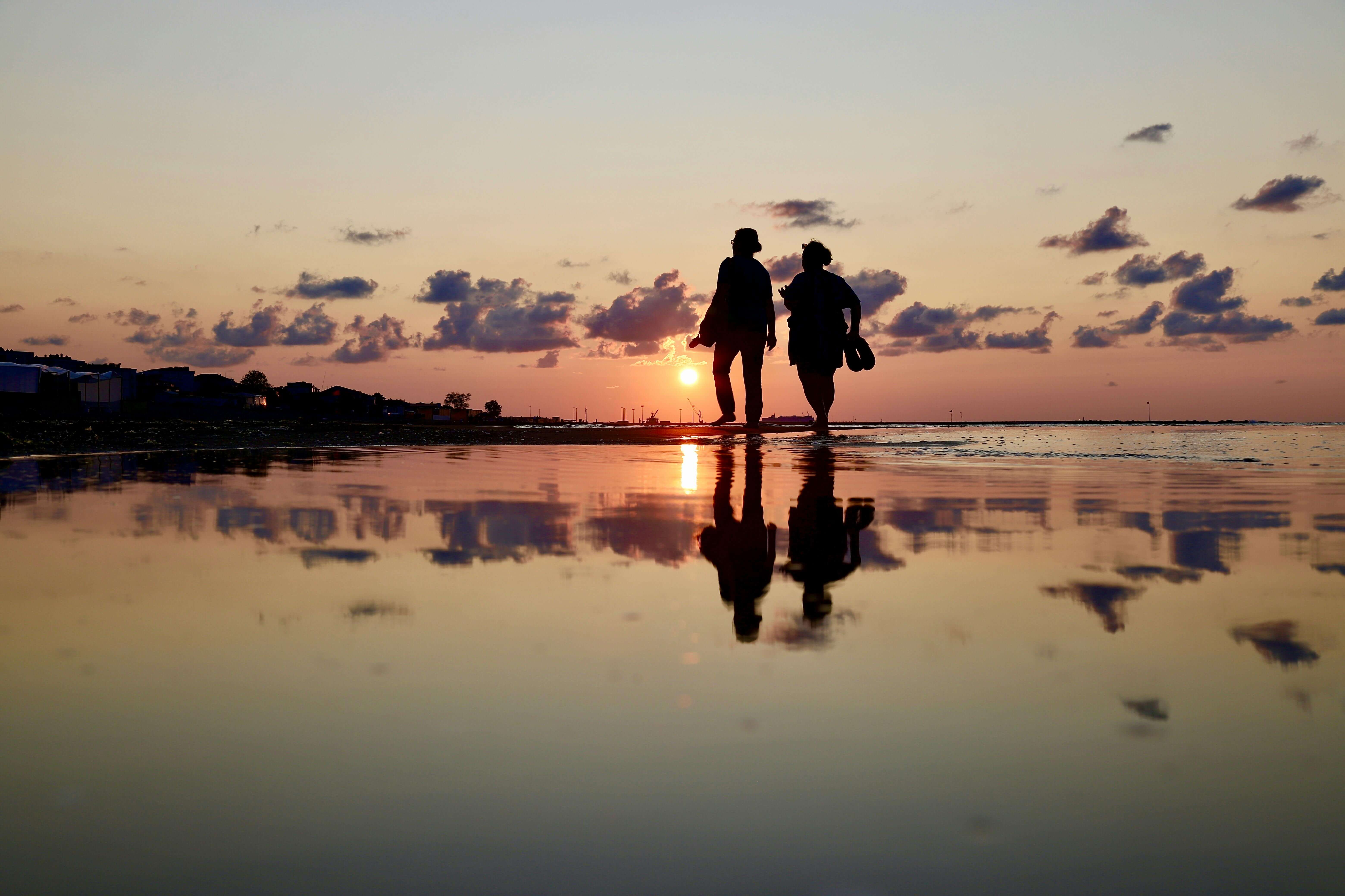 Two individuals walk together at sunset along a reflective shoreline, creating a serene scene.
