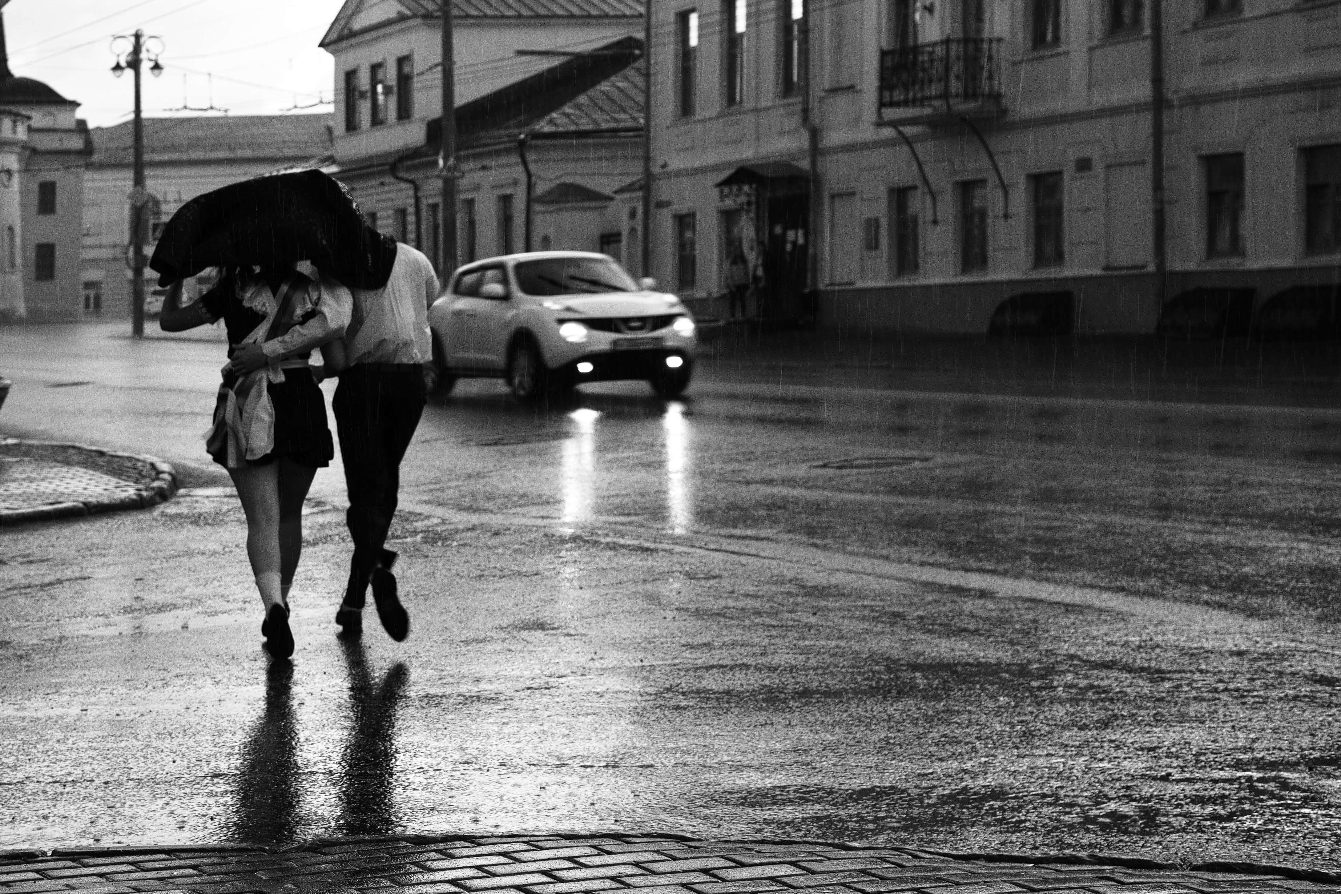 A couple sharing an umbrella cross a wet street as a car approaches in the rain.