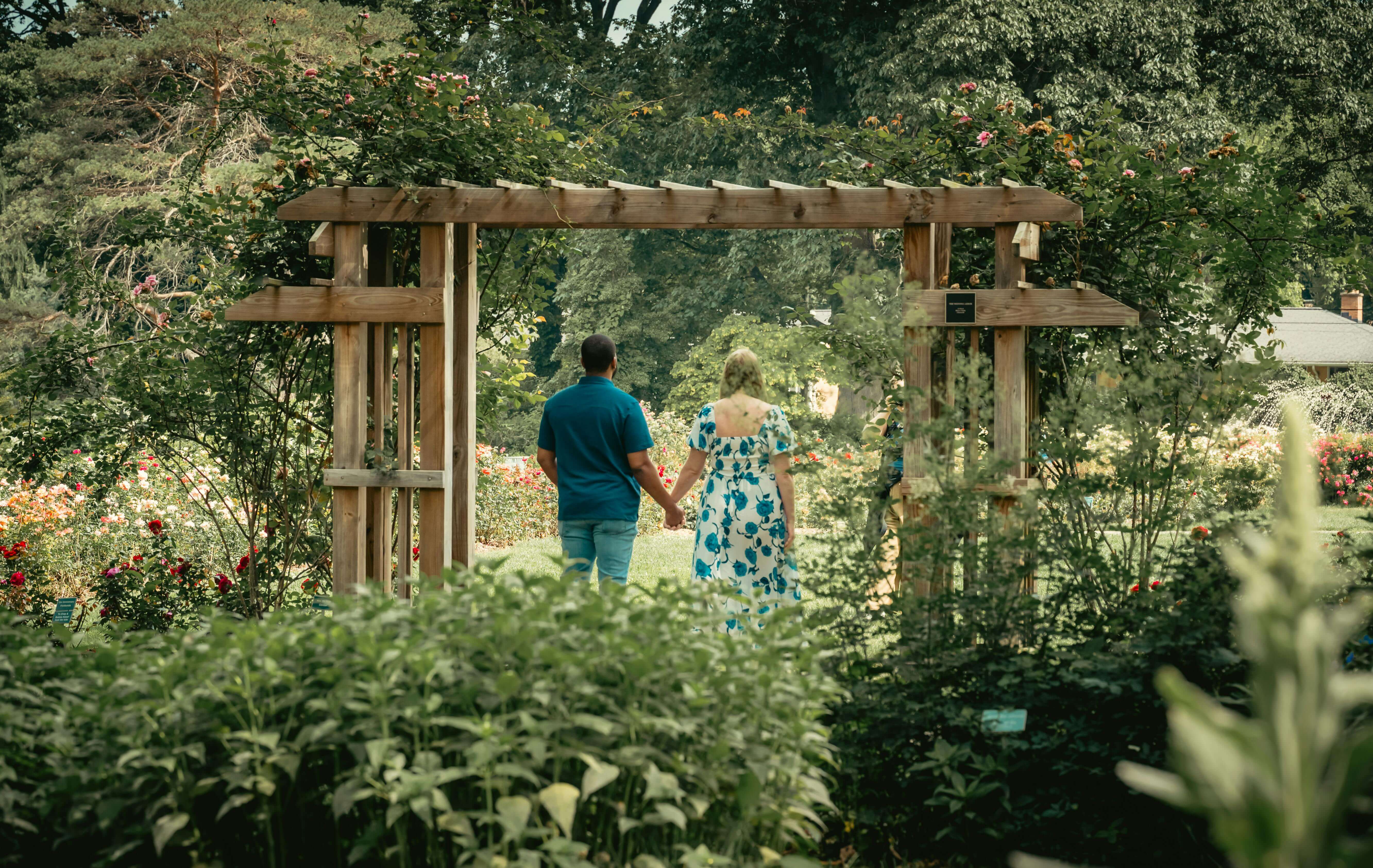 Romantic couple holding hands and walking through a beautiful garden archway, surrounded by lush greenery and vibrant flowers.