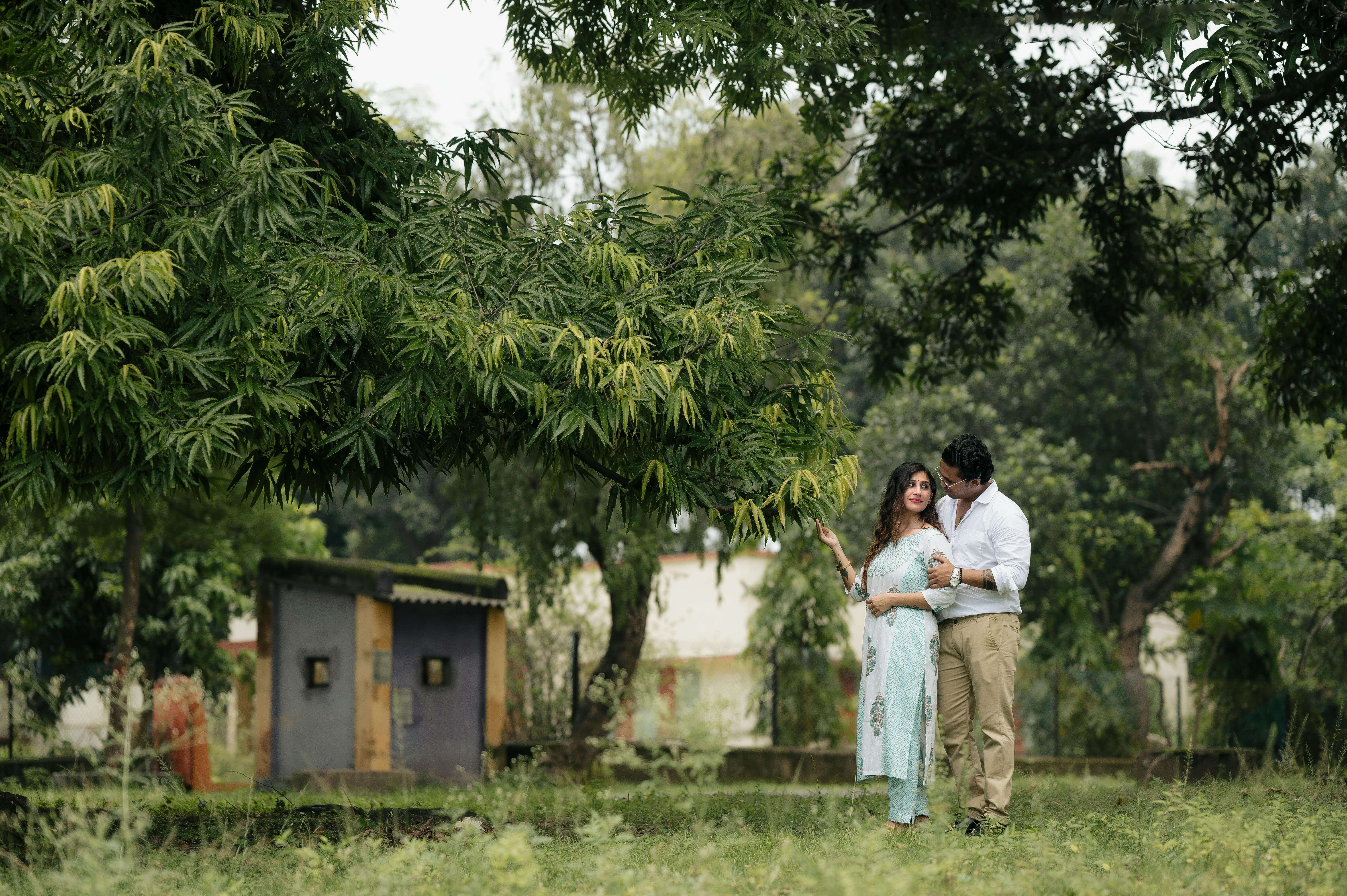 A couple enjoys a romantic moment in a lush green park under a large tree.